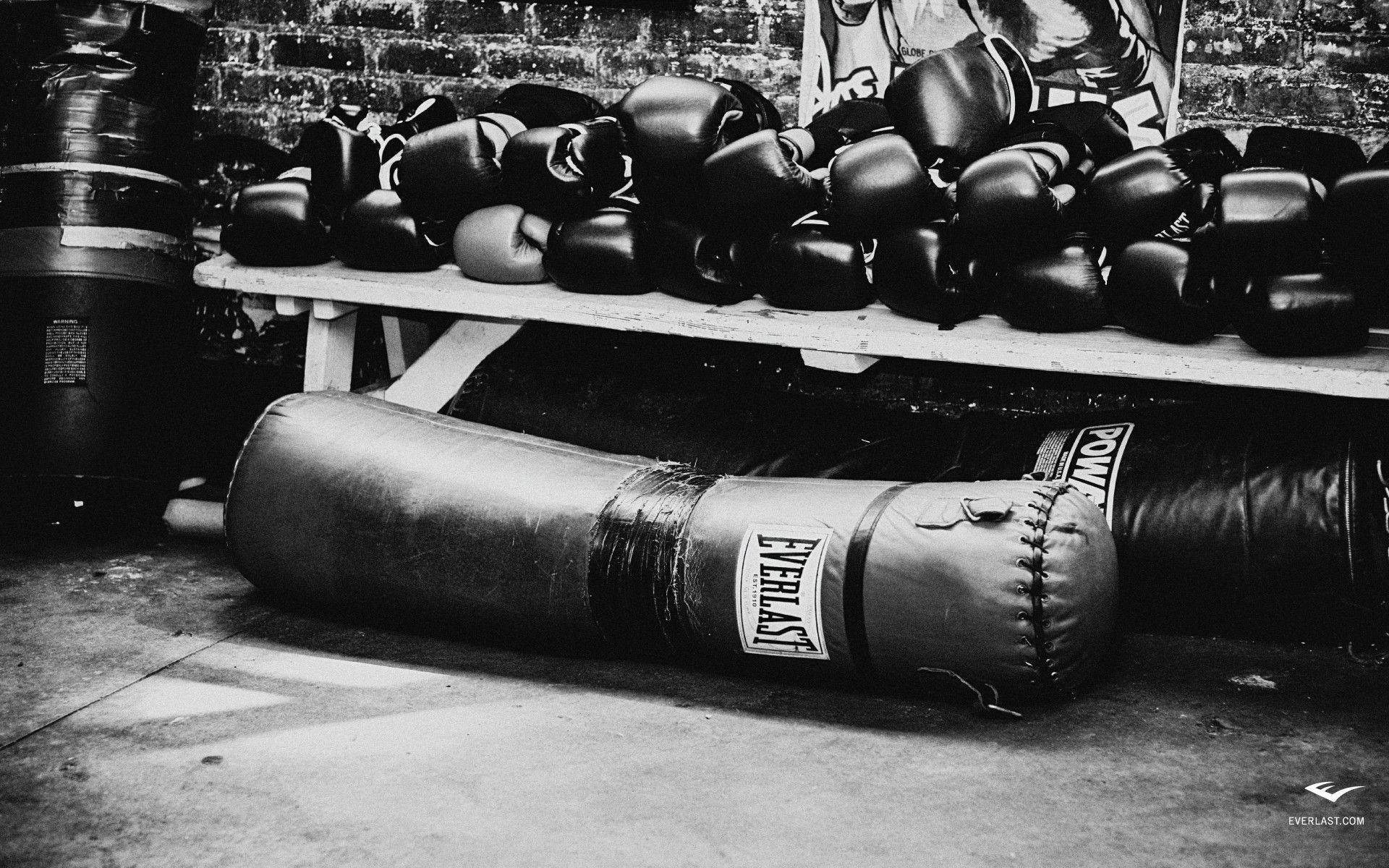 Boxing 桌布 壁紙 — 一張黑白照片，拍攝於一個簡陋的健身房，拳擊手套堆疊在長凳上，旁邊有一個倒下的Everlast沙包。