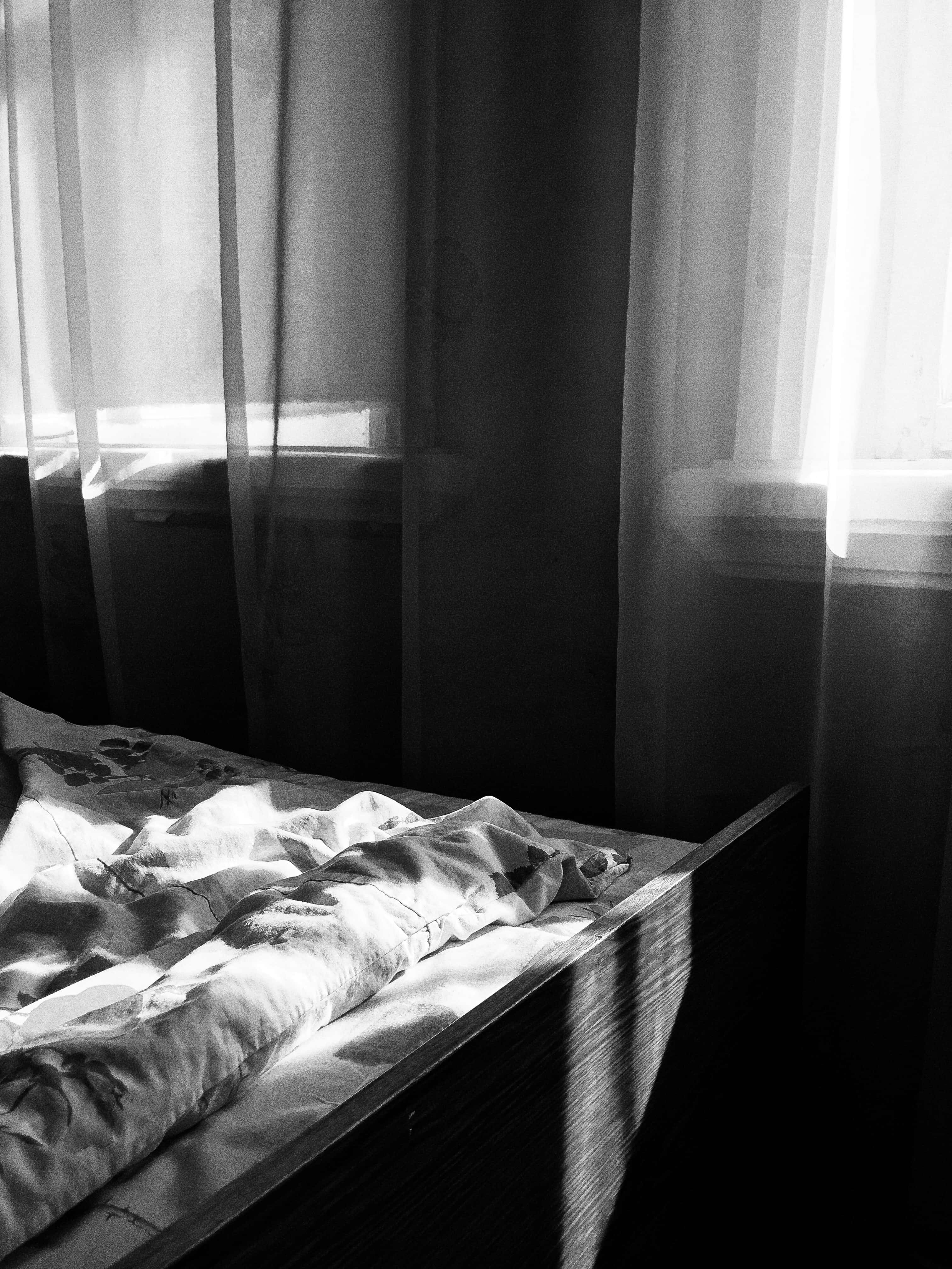Aesthetic Black And White 桌布 壁紙 — 一張黑白照片，拍攝昏暗房間中一張凌亂的床，陽光透過薄紗窗簾照射進來。