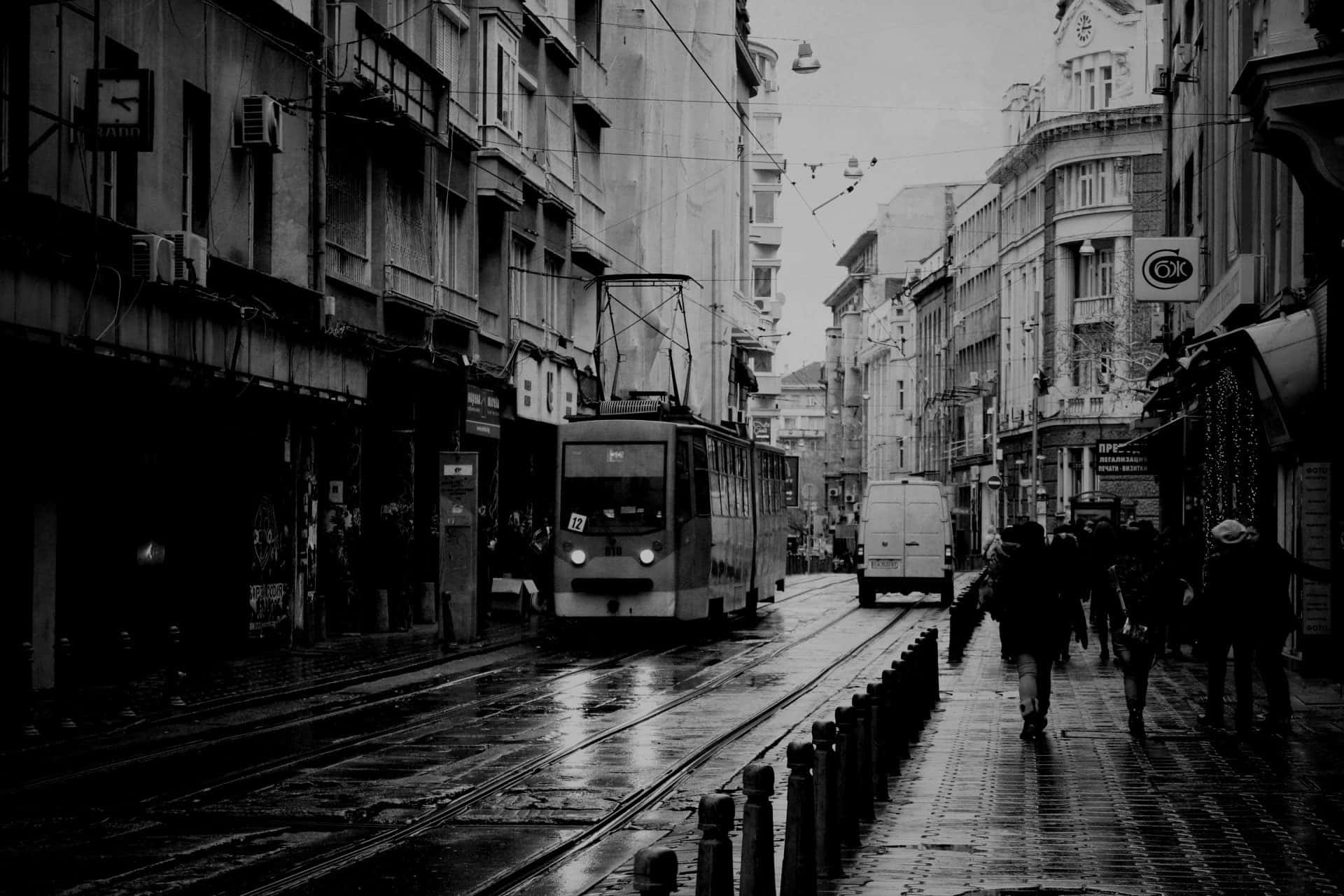Black And White City 桌布 壁紙 — 一張黑白照片，拍攝雨中的城市街道，有電車和行人在濕漉漉的鵝卵石上行走。