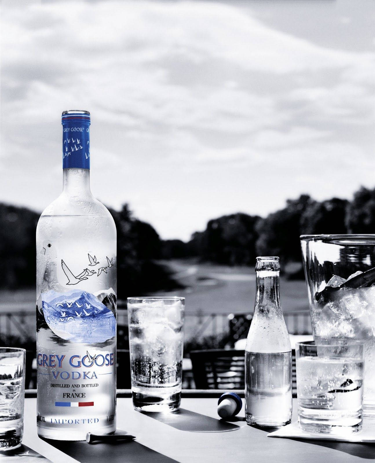 alcohol 桌布 壁紙 — 一張黑白照片，桌上放著 Grey Goose 伏特加酒瓶和酒杯，背景是模糊的戶外景色。