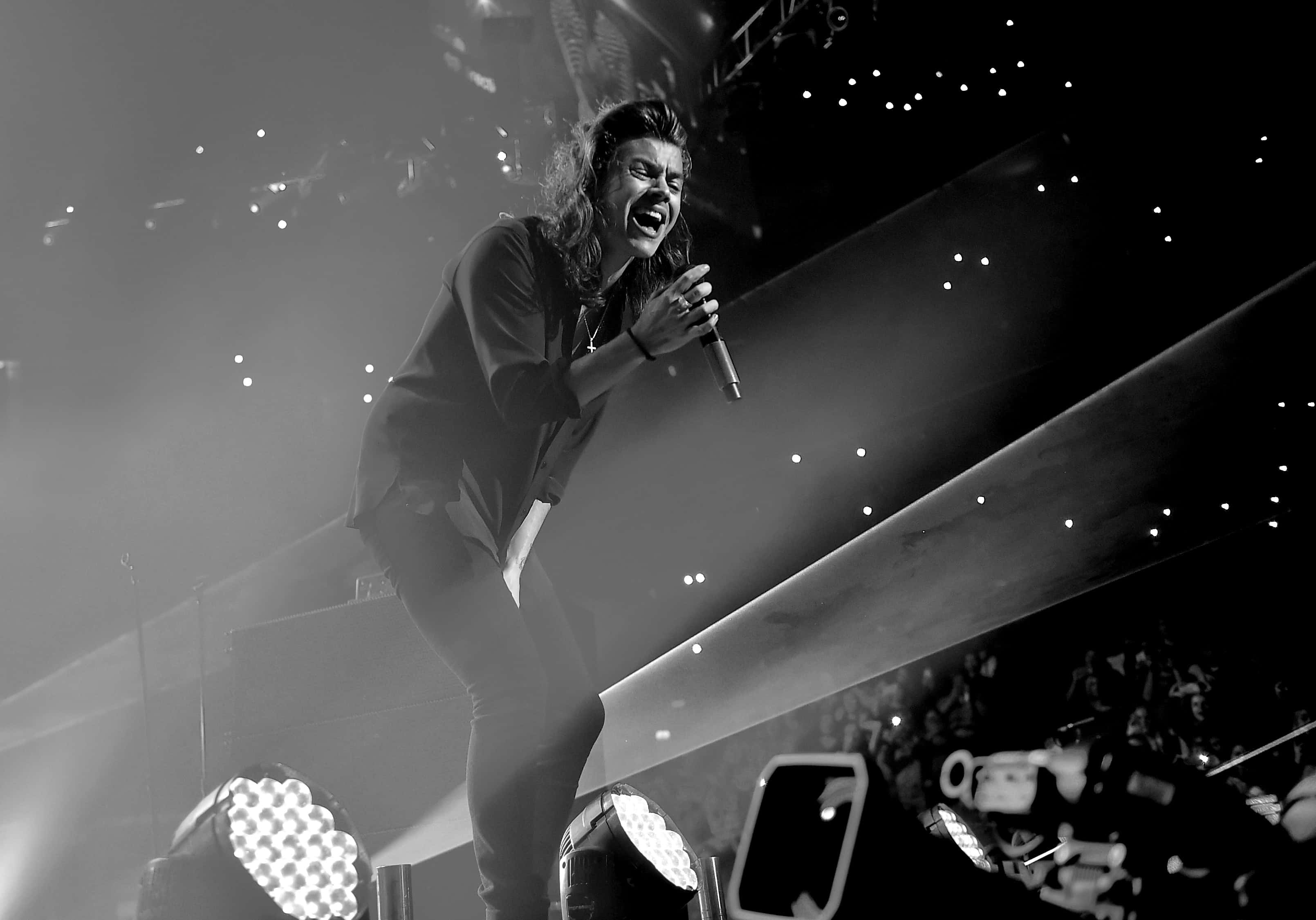 Harry Styles Black And White 桌布 壁紙 — 一張黑白照片，歌手在舞台上充滿激情地表演，背景有聚光燈和人群。