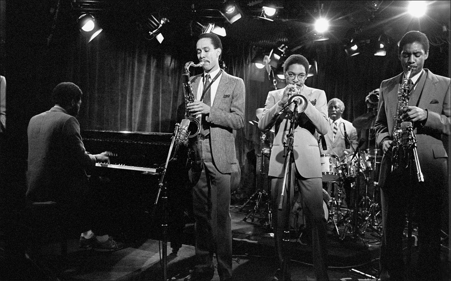 Jazz 桌布 壁紙 — 一張黑白照片，爵士樂隊在明亮的聚光燈下在舞台上表演，鋼琴家和薩克斯風手身穿西裝。