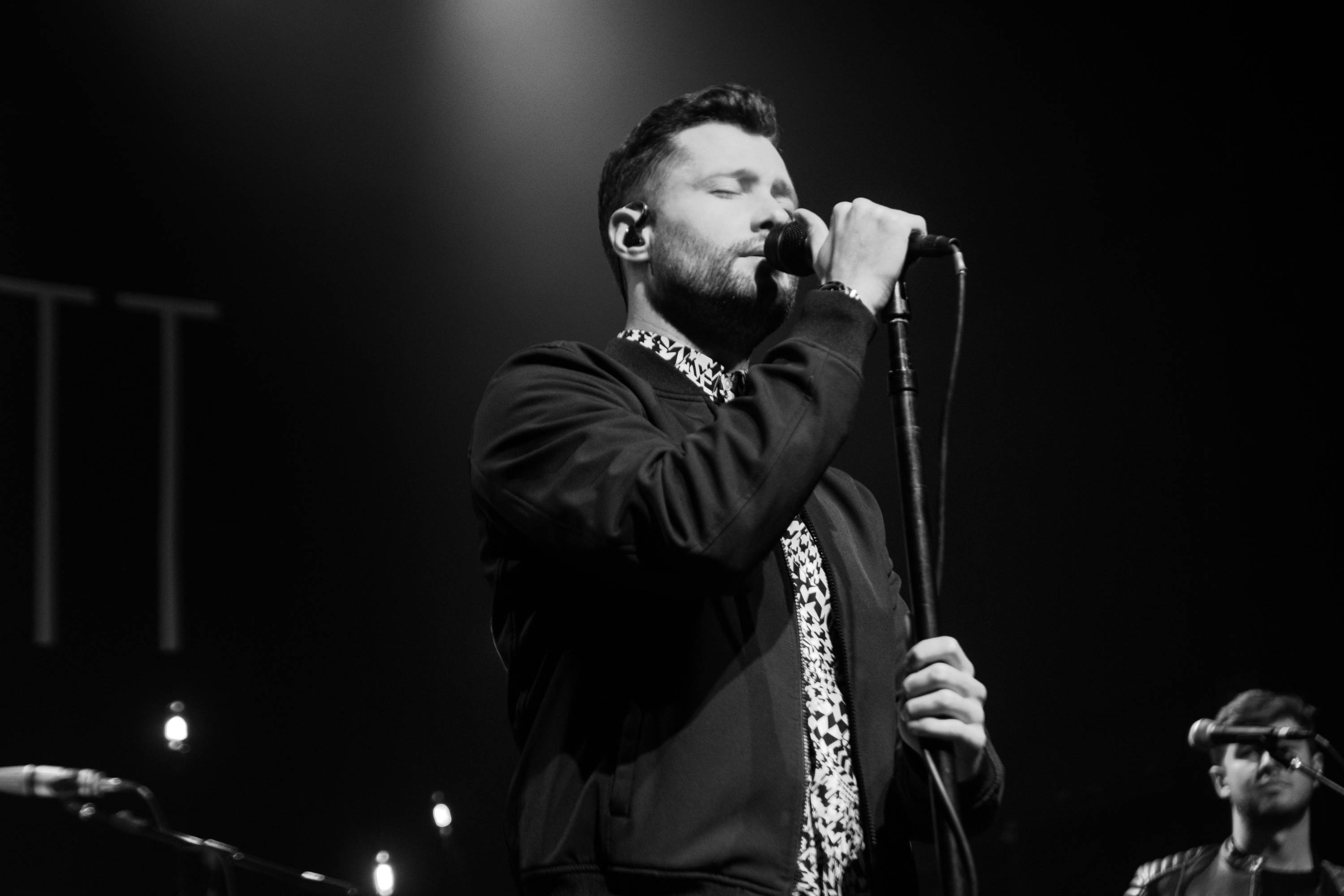 Calum Scott 桌布 壁紙 — 一張黑白照片，顯示一位男歌手在聚光燈下熱情地在舞台上表演，背景中可見一位樂隊成員。