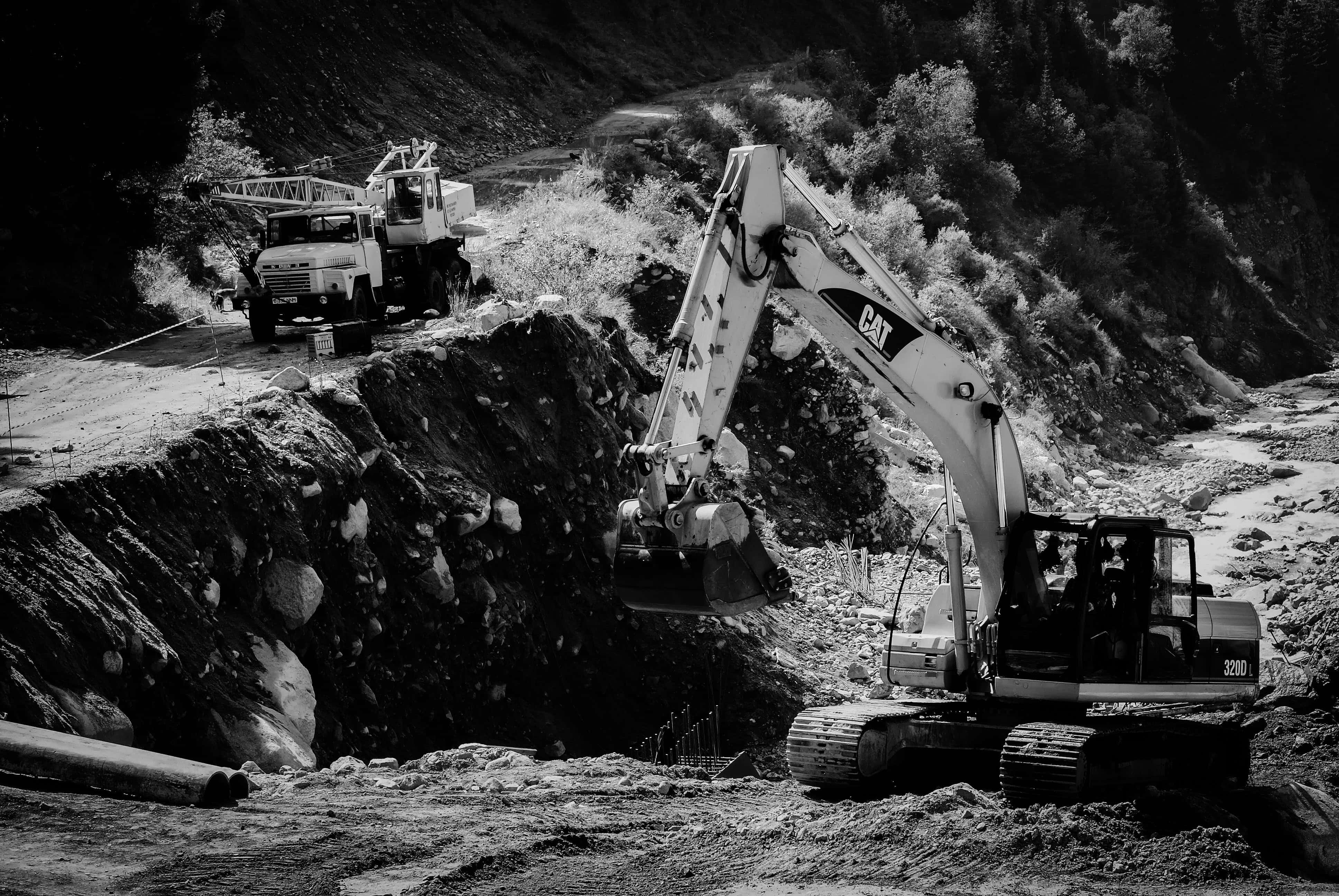 Mining 桌布 壁紙 — 一張黑白照片，顯示一輛CAT挖土機正在停放的工程車旁邊的岩石山坡上挖掘。