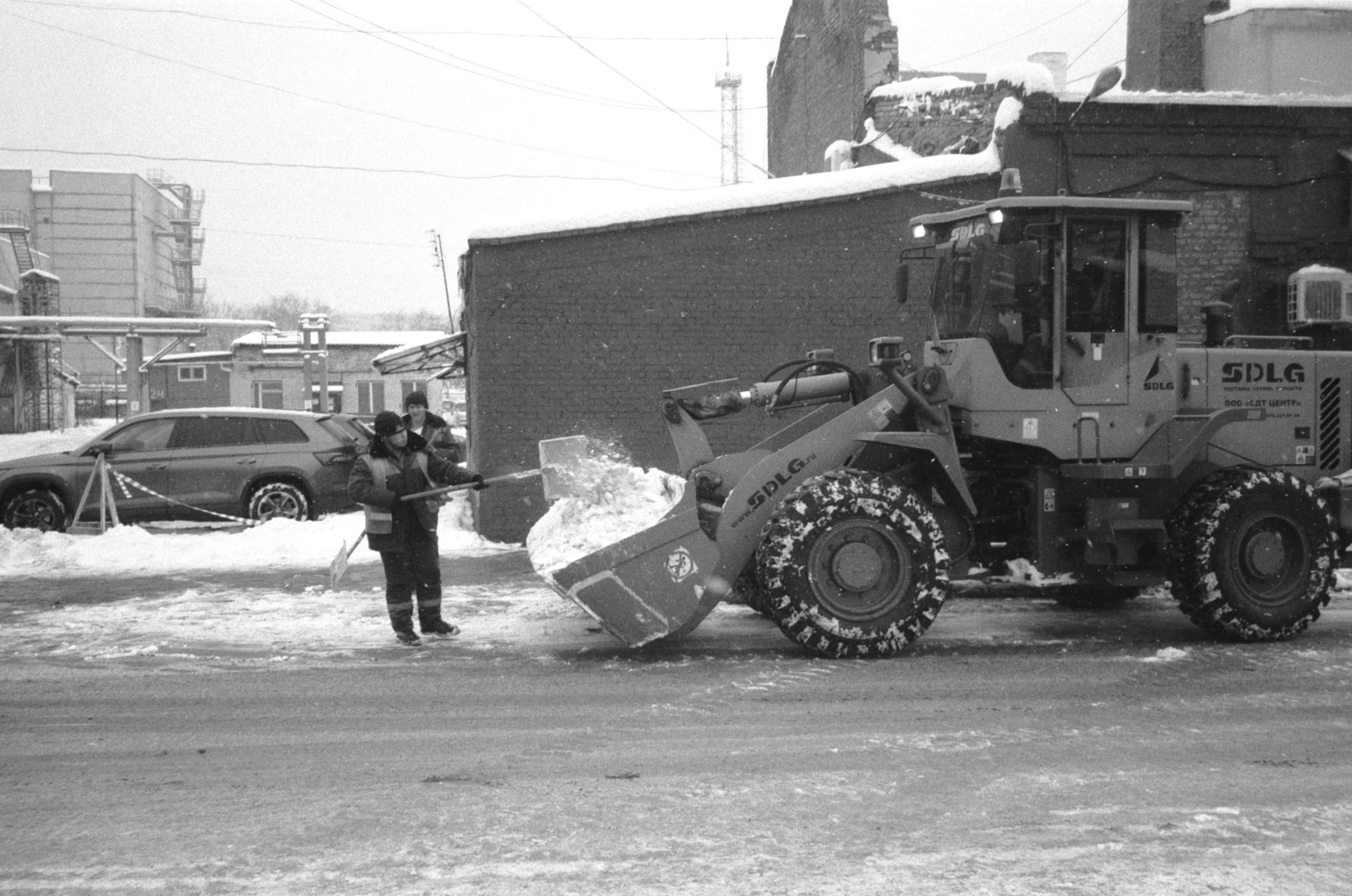Snowplow 桌布 壁紙 — 一張黑白照片，顯示一輛掃雪車正在清理街道上的積雪，而兩名工人在附近剷雪，背景是雪白的城市環境。