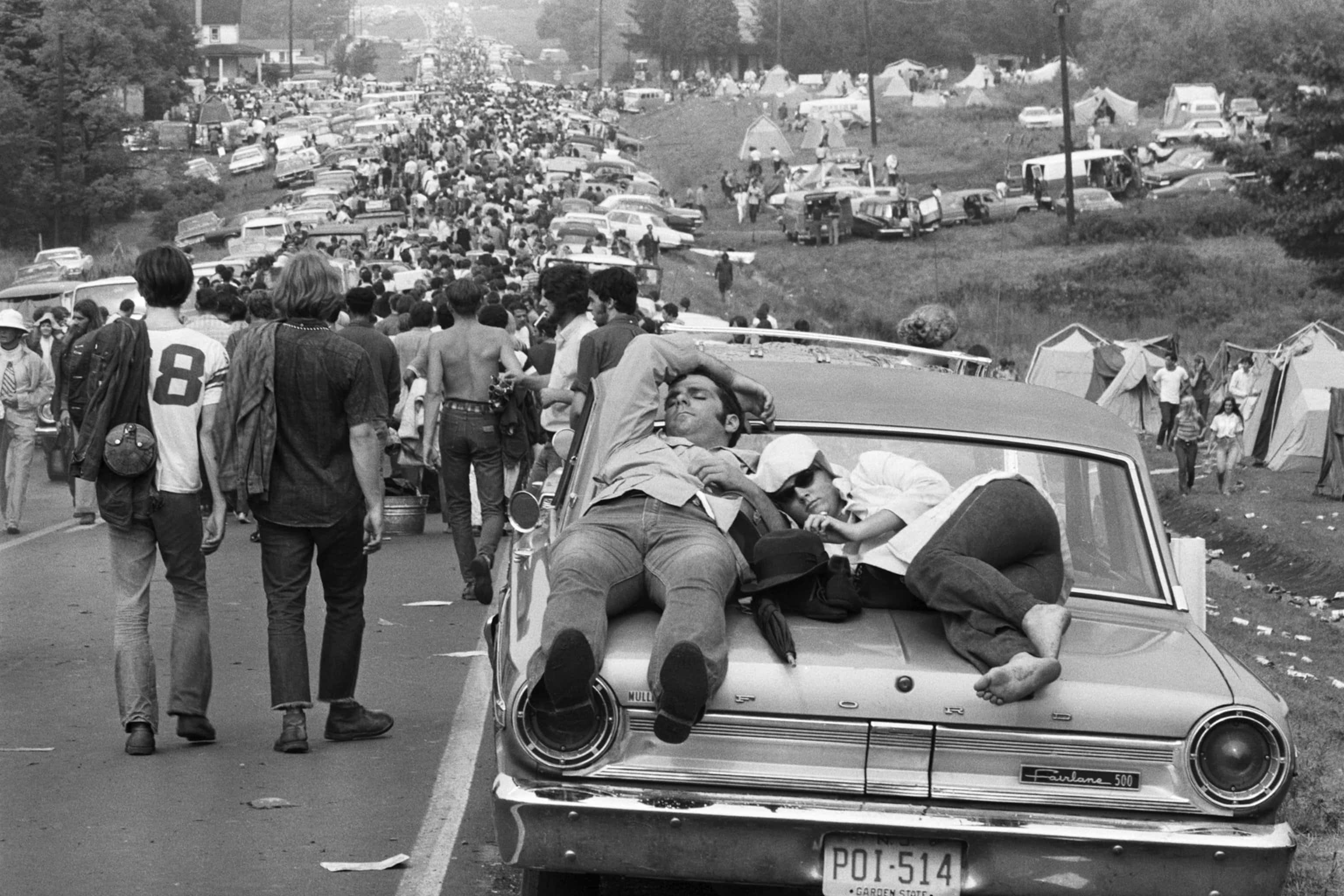 woodstock 桌布 壁紙 — 一張黑白照片捕捉了1960年代音樂節的人群，人們在經典汽車引擎蓋上休息，而其他人則在停放的車輛和帳篷之間走動。