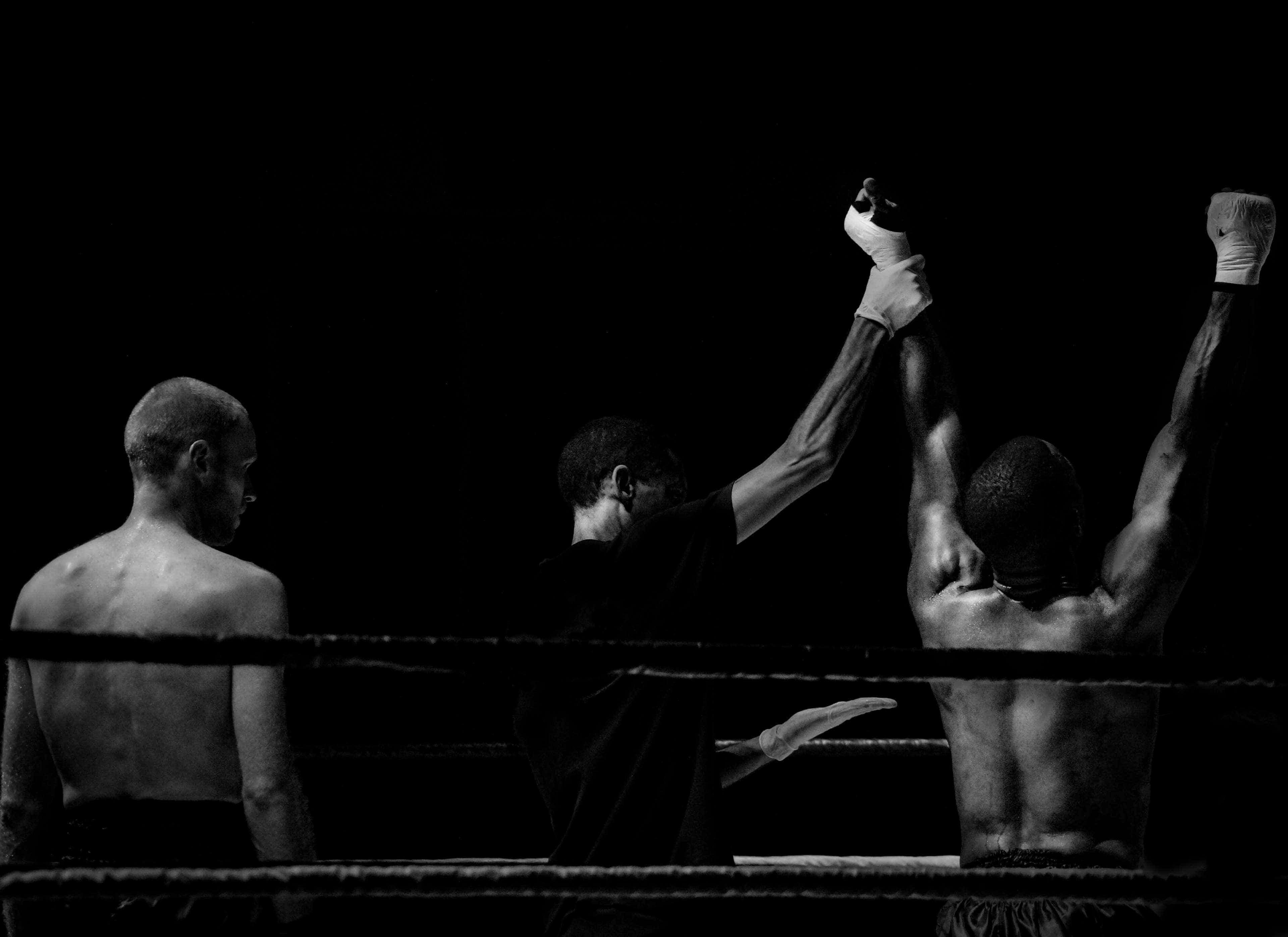 Boxing 桌布 壁紙 — 一張黑白照片捕捉到一位勝利的拳擊手高舉雙臂，裁判在昏暗的擂台上宣布勝利。