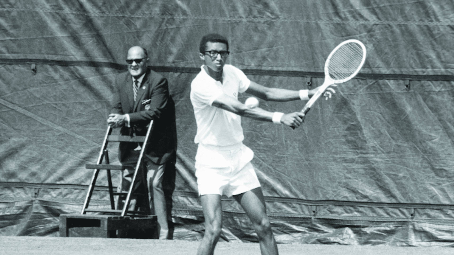 Tennis 桌布 壁紙 — 一張黑白照片捕捉到一名網球選手正在揮拍，球在運動中，而一名穿西裝的男子從梯子上觀看。