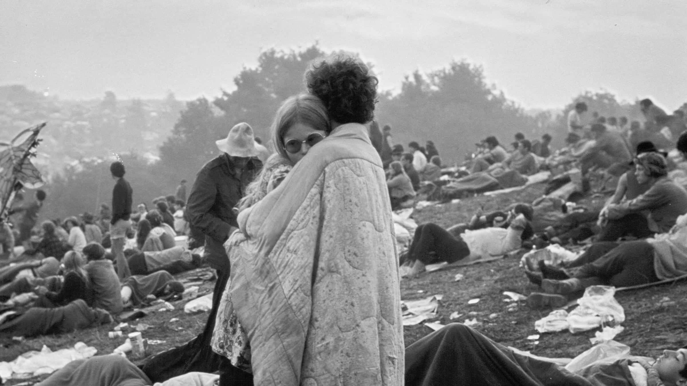 Woodstock 桌布 壁紙 — 一張黑白照片捕捉到一對情侶在長滿草的山坡上擁抱，背景是擁擠的戶外聚會，人們在休息。