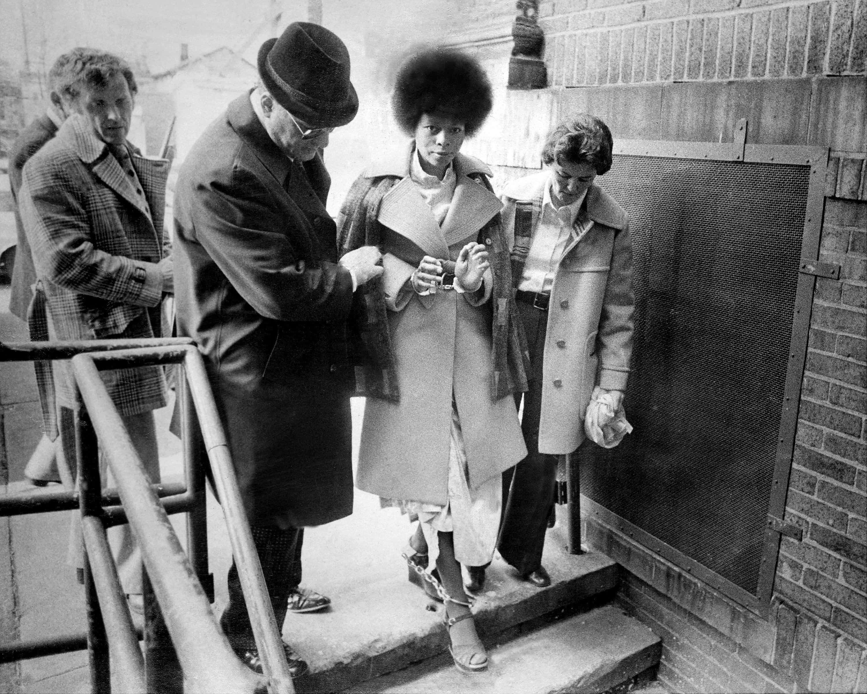 Black Panther Party 桌布 壁紙 — 一張黑白照片顯示一名有非洲式髮型的女子被兩名穿著大衣的男子護送下戶外樓梯。