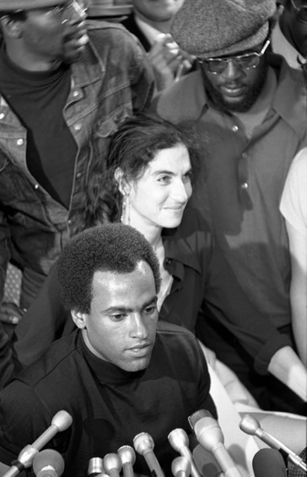 Black Panther Party 桌布 壁紙 — 一張黑白照片顯示一名男子在記者會上發言，周圍佈滿麥克風，身後的人們專心聆聽。