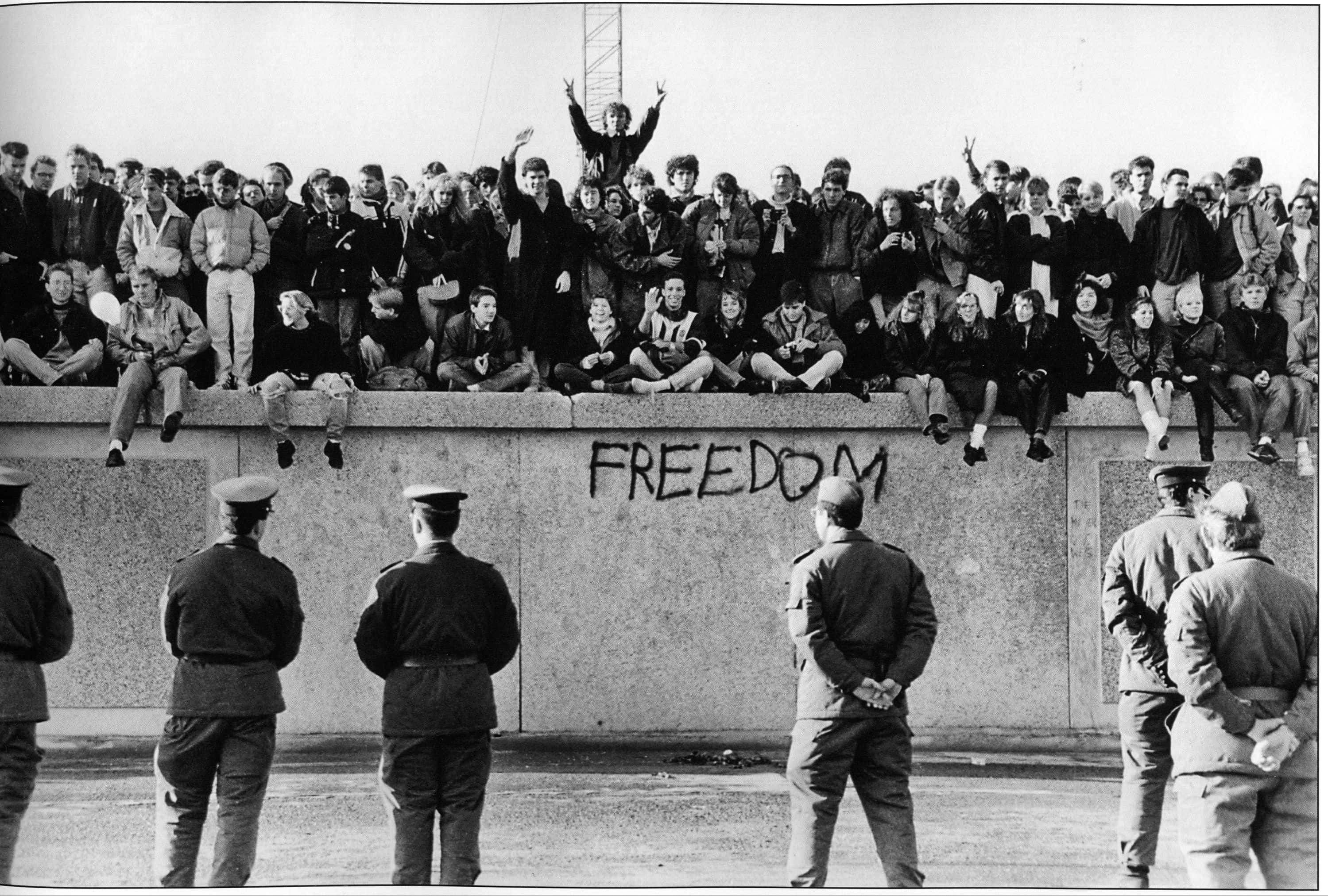 wall 桌布 壁紙 — 一張黑白照片顯示一群人坐在混凝土牆上，牆上噴有「FREEDOM」字樣，而身穿制服的警衛站在下方。