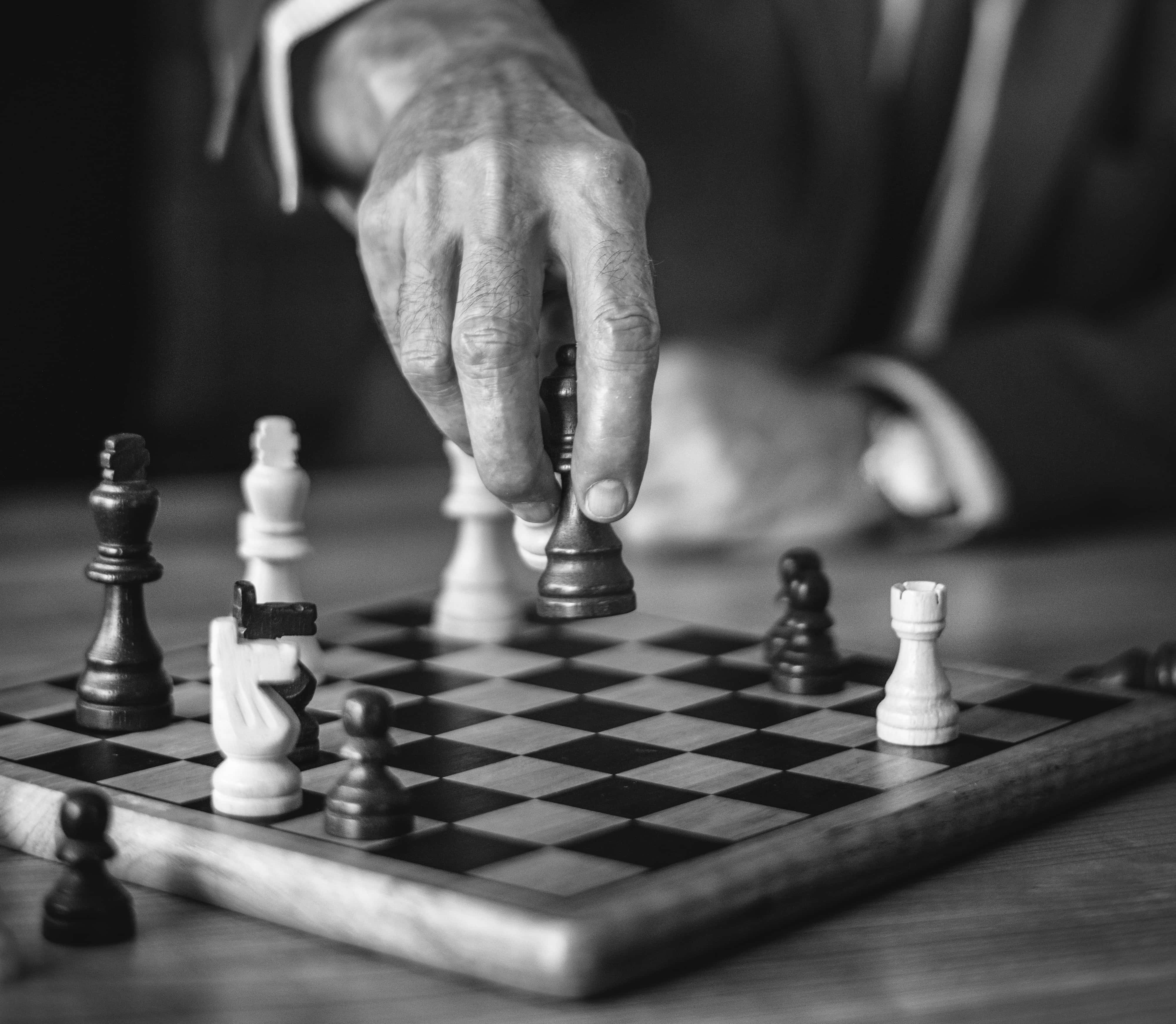 Black And White Chess 桌布 壁紙 — 一張黑白特寫，一隻飽經風霜的手將一枚棋子舉起，下方木製棋盤上還有其他棋子正在對弈。