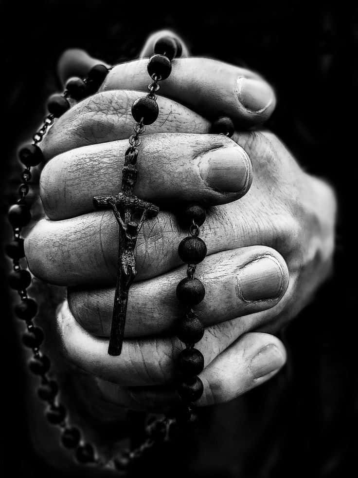 Praying 桌布 壁紙 — 一張黑白特寫照片，展示粗糙的雙手緊緊握著帶有十字架吊墜的玫瑰經念珠。
