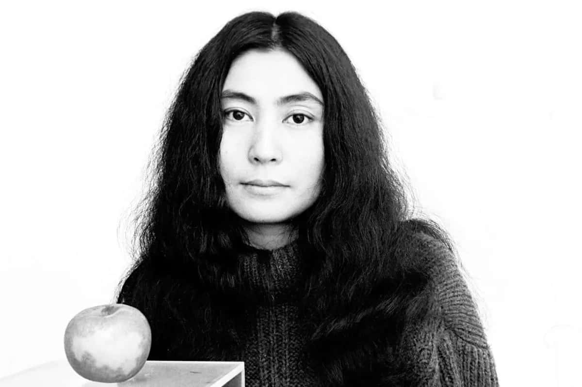 serious face 桌布 壁紙 — 一張黑白肖像照，一位長黑髮的女性在桌上一個蘋果旁凝視前方。