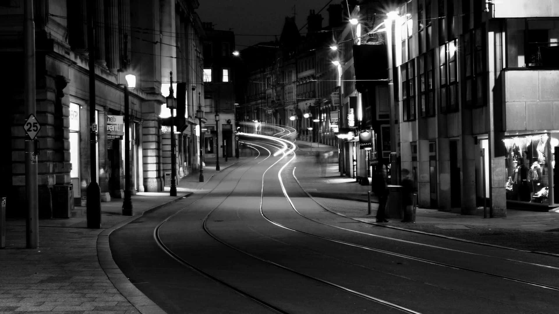 Black And White Street 桌布 壁紙 — 一張黑白長曝光照片，拍攝夜晚彎曲的城市街道，帶有發光的電車燈軌跡和燈火通明的建築物。