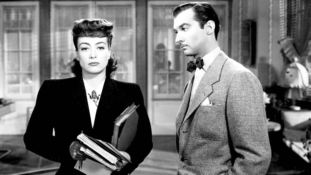 Film Noir 桌布 壁紙 — 一張黑白電影劇照顯示一位身穿深色西裝的女性拿著文件，站在一位身穿格紋西裝和領結的男性旁邊，室內場景。