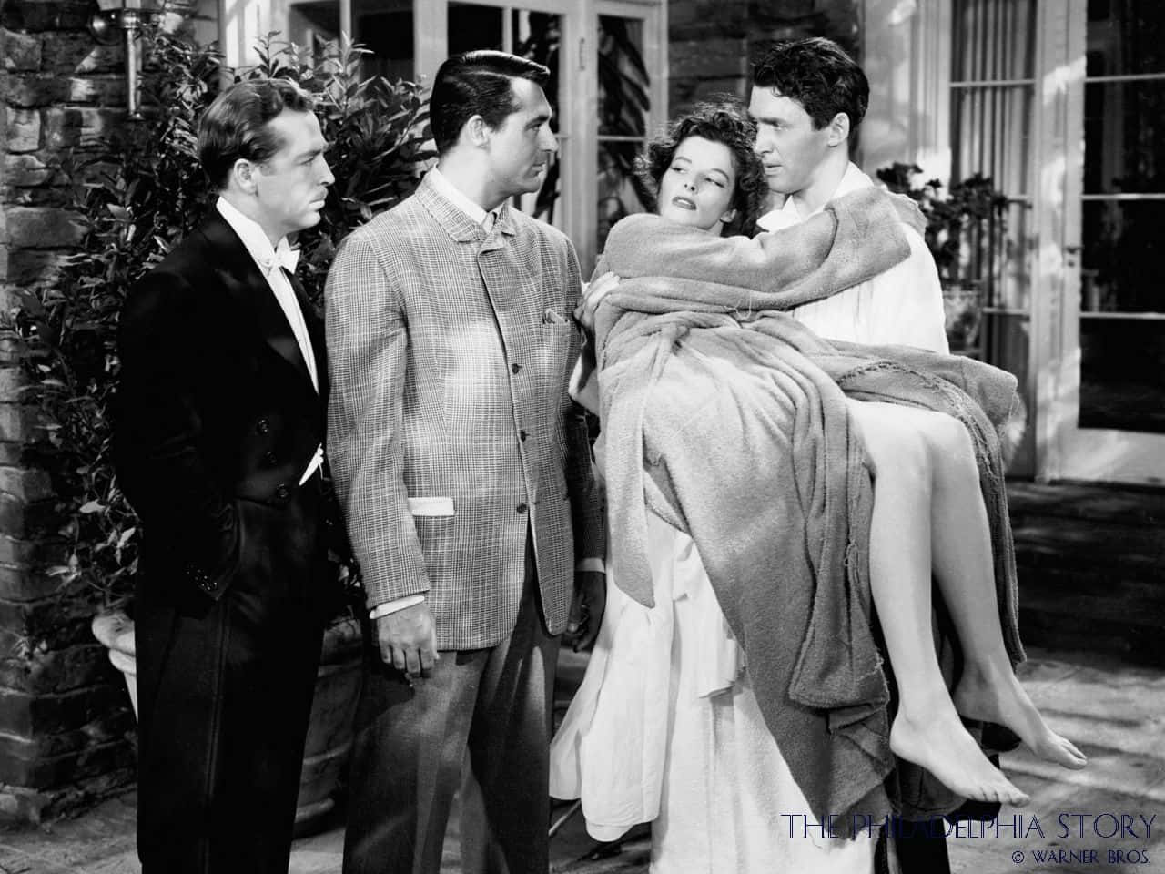 Classic Movie 桌布 壁紙 — 一張黑白電影劇照顯示一名男子抱著一名裹著毯子的女子，而另外兩名男子則在戶外專注地觀看。