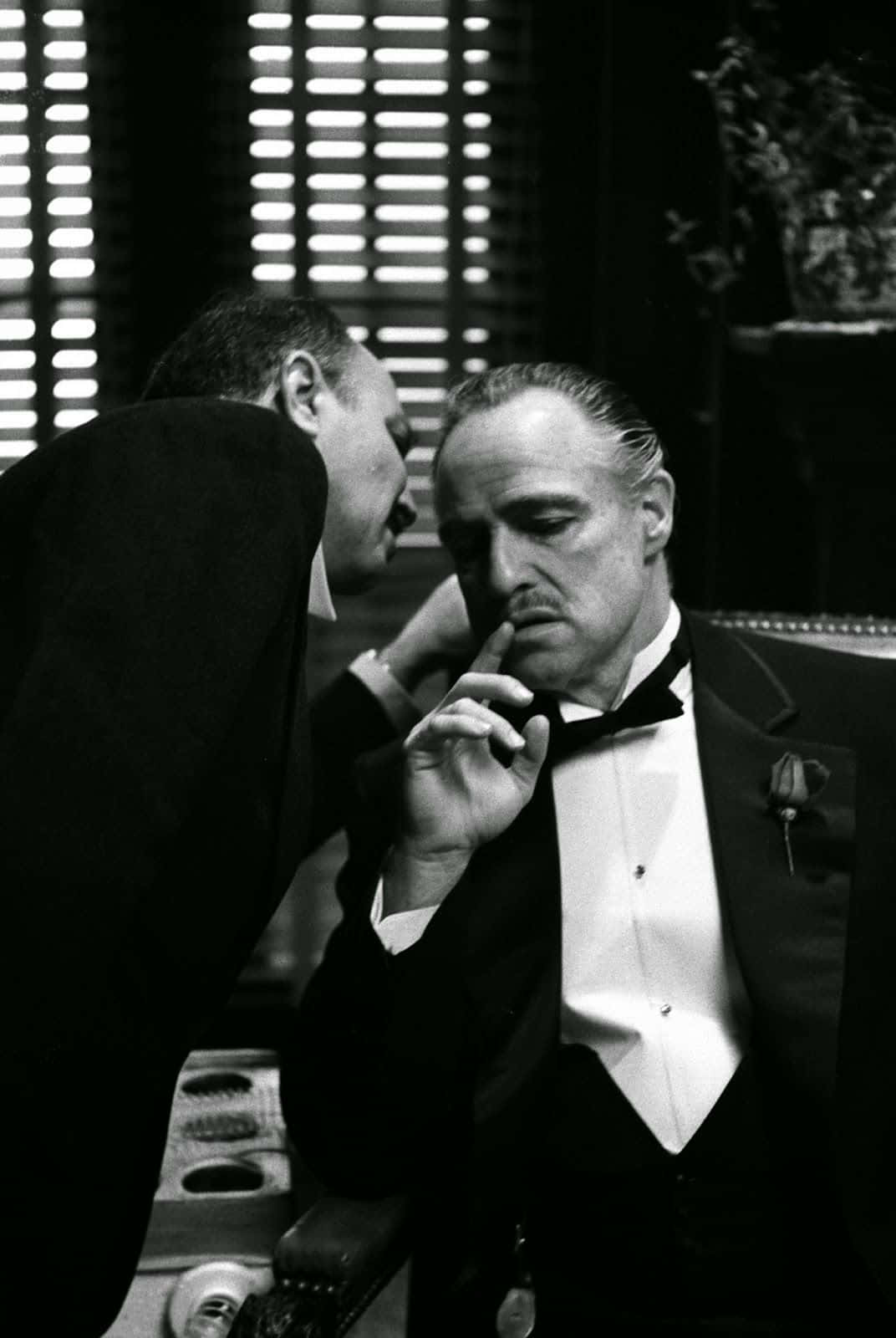 Michael Corleone 桌布 壁紙 — 一張黑白電影劇照顯示一名身穿燕尾服的男人坐著，另一名男人則靠近他，營造出緊張而親密的時刻。