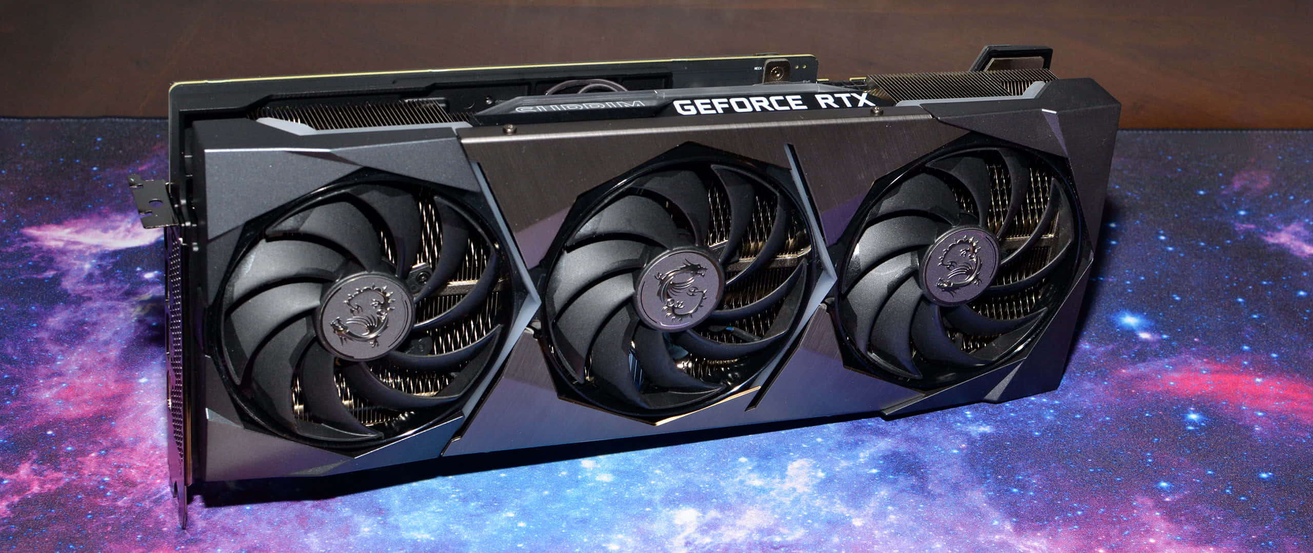 msi 2560x1080 桌布 壁紙 — 一張黑色MSI GeForce RTX顯示卡，配備三個風扇，放置在一個帶有紫色、藍色和粉紅色星星的宇宙星雲桌布上。