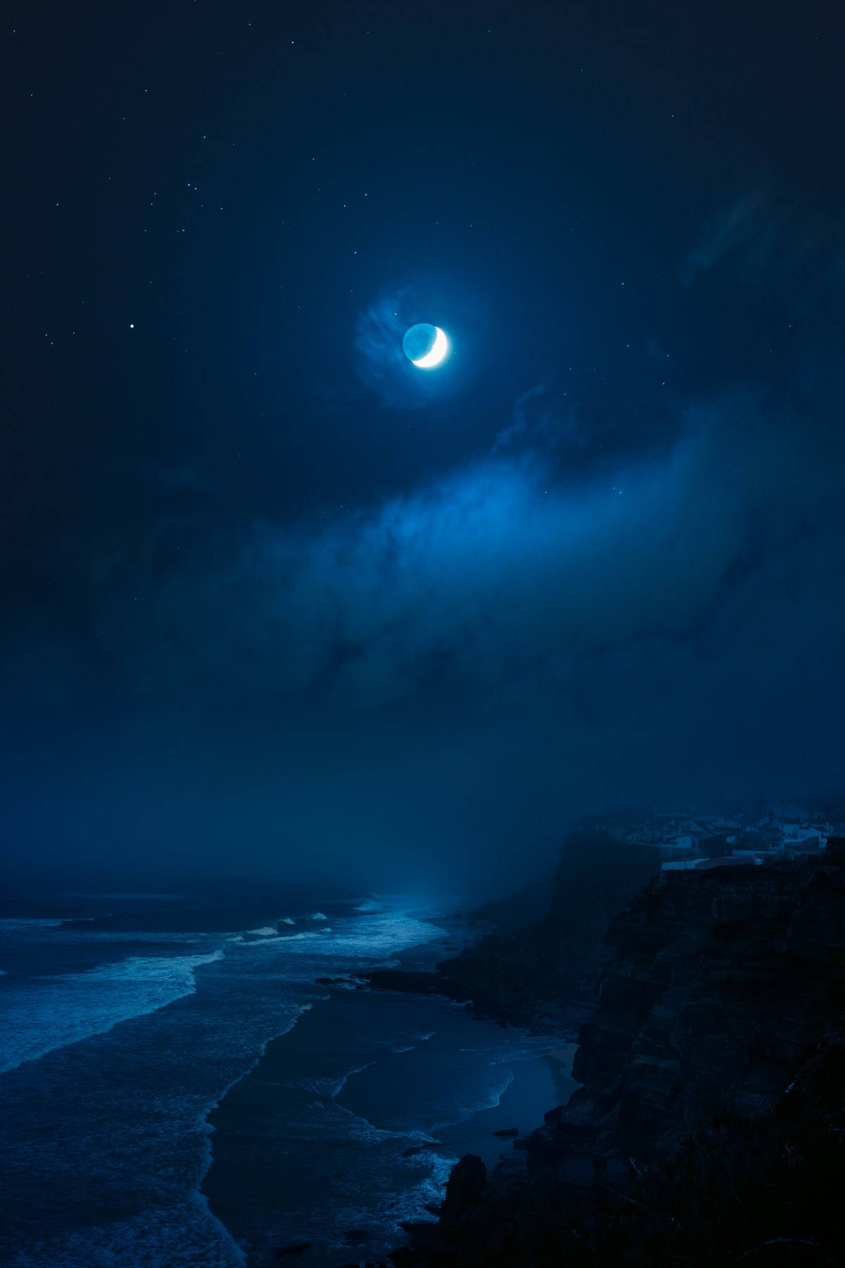 iPhone 桌布 壁紙 — 一彎新月在繁星點點的夜空下，透過輕柔的雲層照耀著黑暗的海洋，海浪拍打著崎嶇的懸崖。