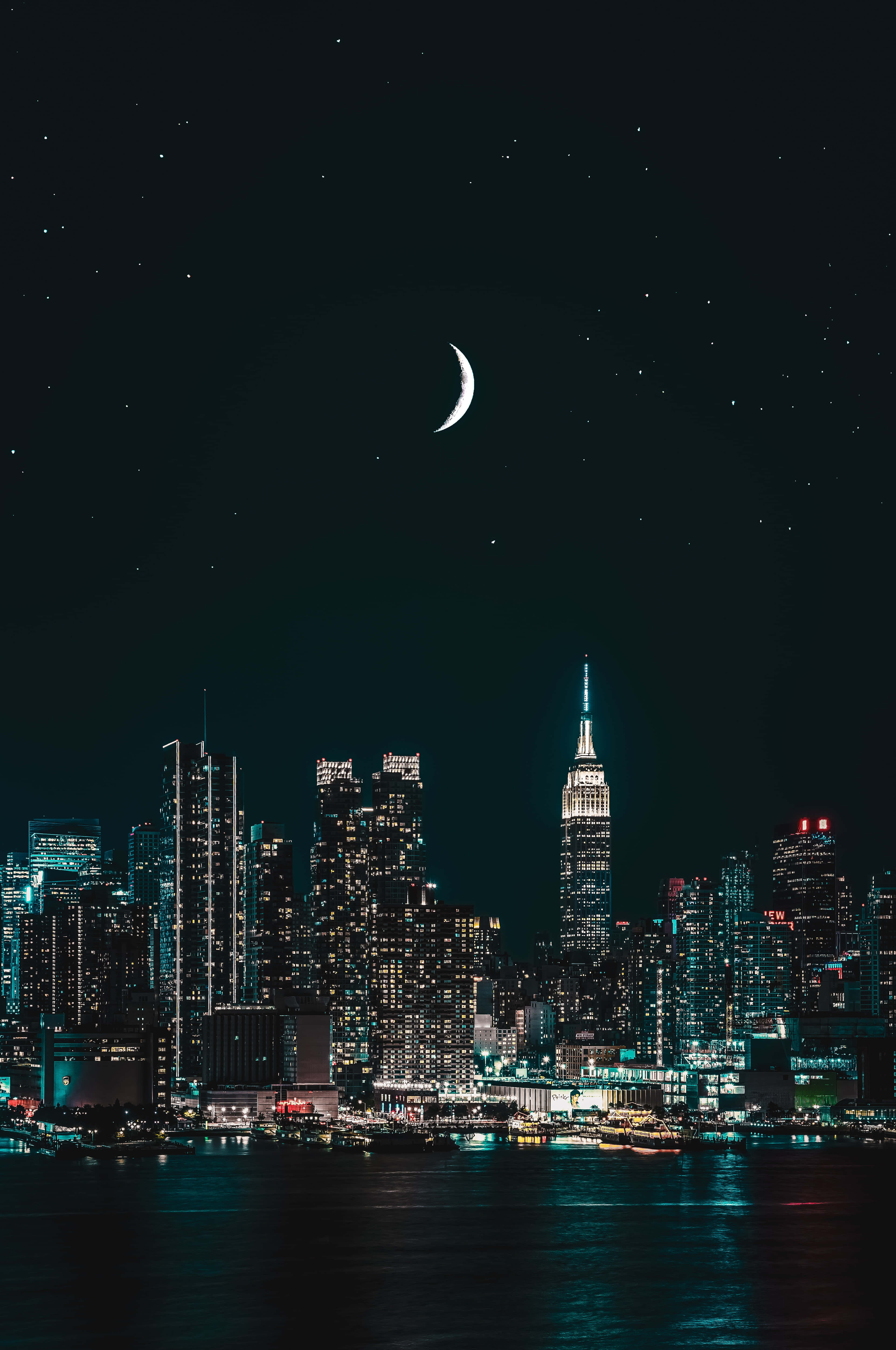 城市風景 桌布 壁紙 — 一彎新月懸掛在閃爍的夜景城市上空，摩天大樓在星空下的暗水中倒映。