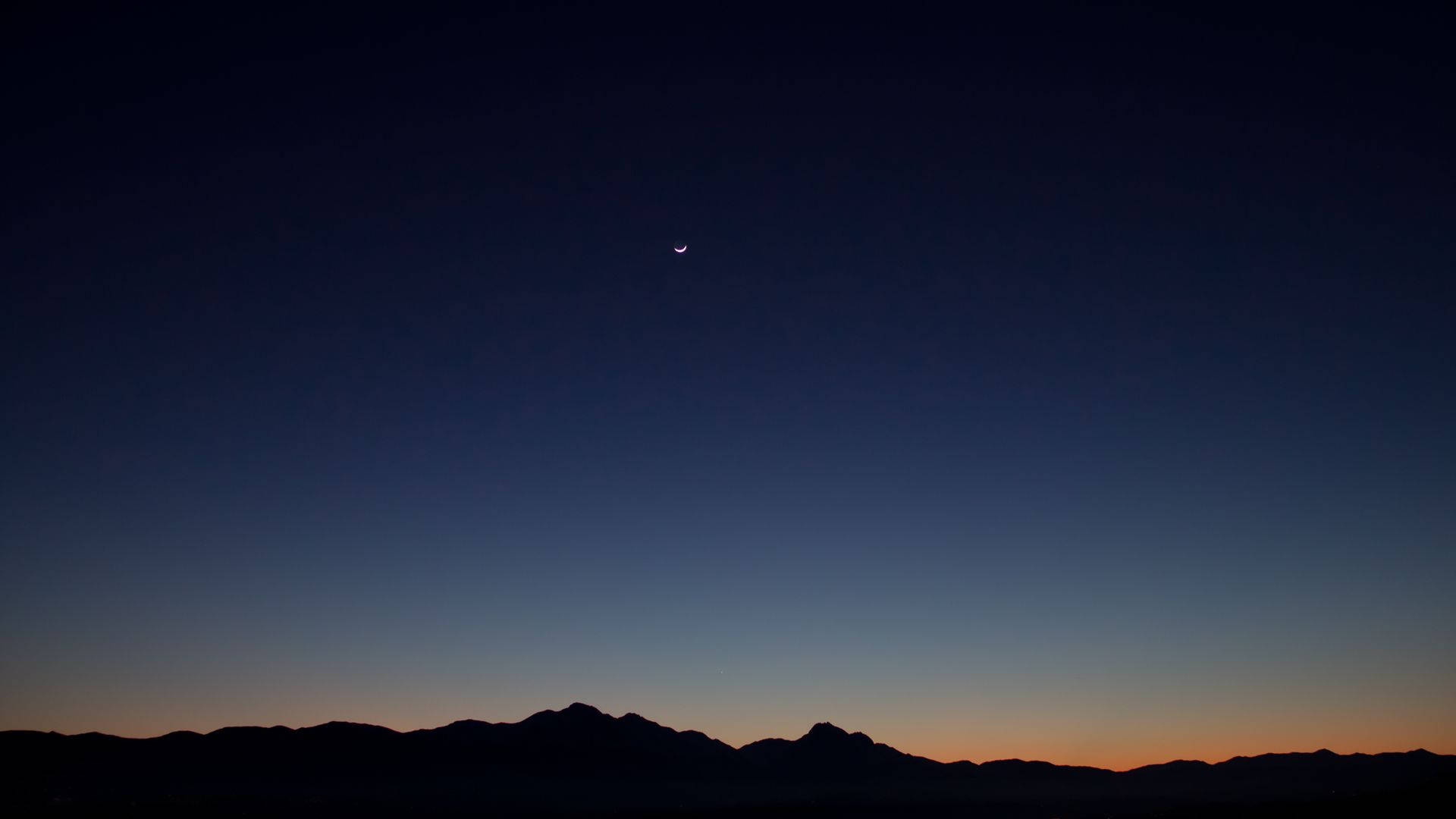 夜空 桌布 壁紙 — 一彎纖細的月牙在暮色天空下照耀著黑暗的山脈，天空從深藍色漸變為地平線上的橙色。