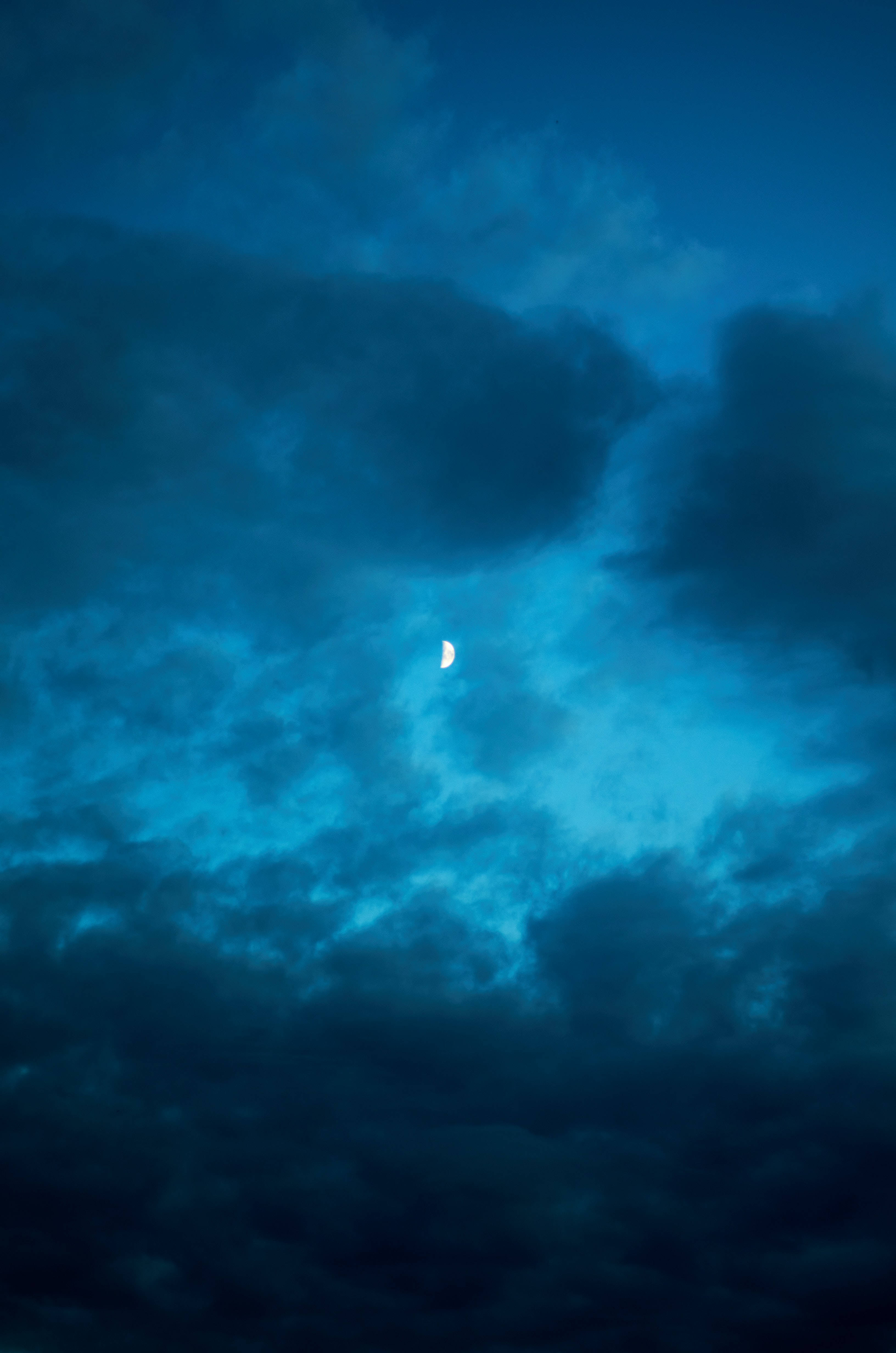 雲朵 桌布 壁紙 — 一彎纖細的月牙在深藍色夜空下，透過旋轉的藍綠色和深藍色雲層柔和發光。