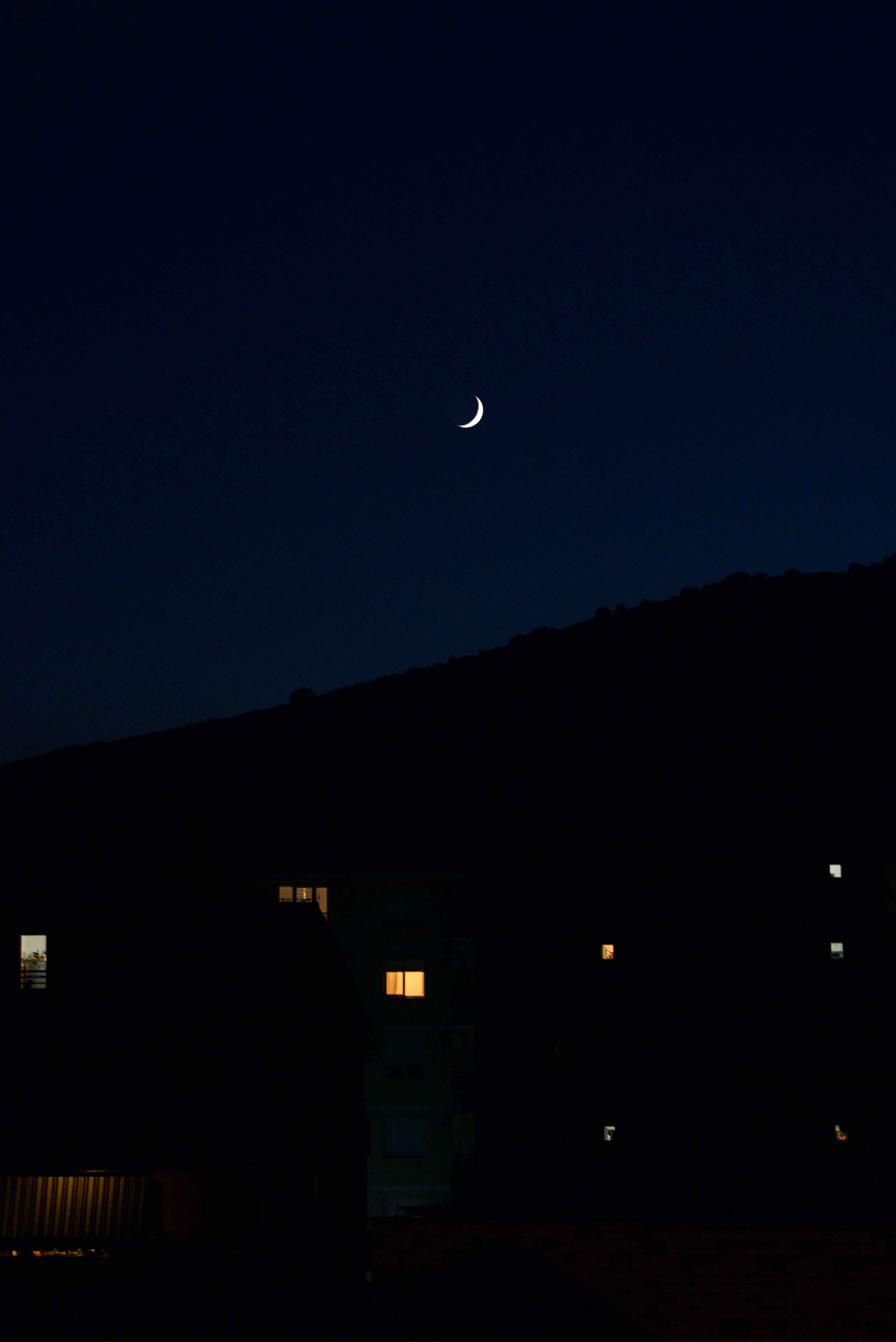 Crescent 桌布 壁紙 — 一彎纖細的月牙在黑暗的山坡上發光，夜間寧靜住宅樓的窗戶裡閃爍著溫暖的燈光。