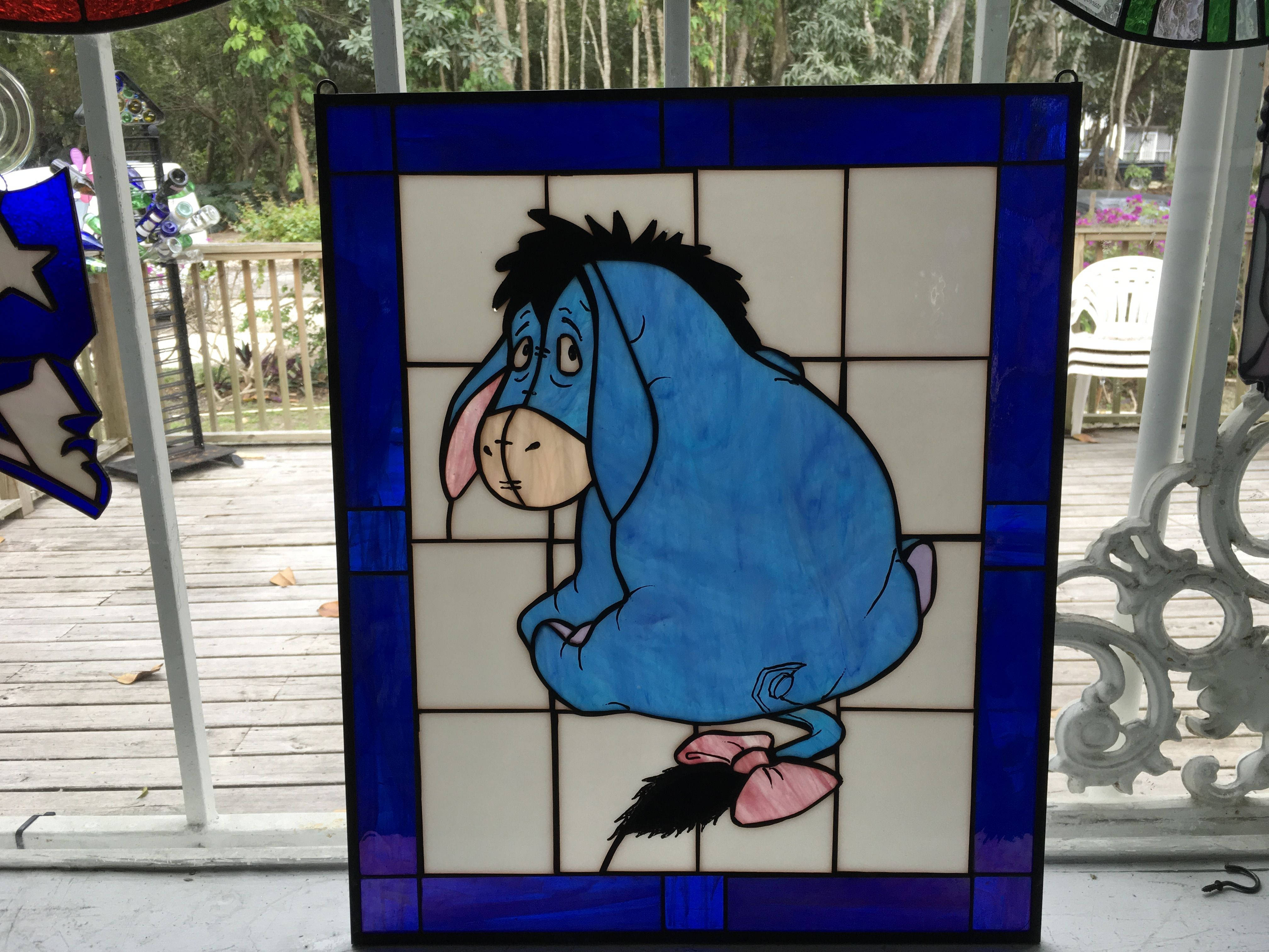 Eeyore 桌布 壁紙 — 一扇彩色玻璃窗，上面有一隻藍色卡通驢子，戴著粉紅色蝴蝶結，位於帶有木質甲板和花園景觀的門廊上。
