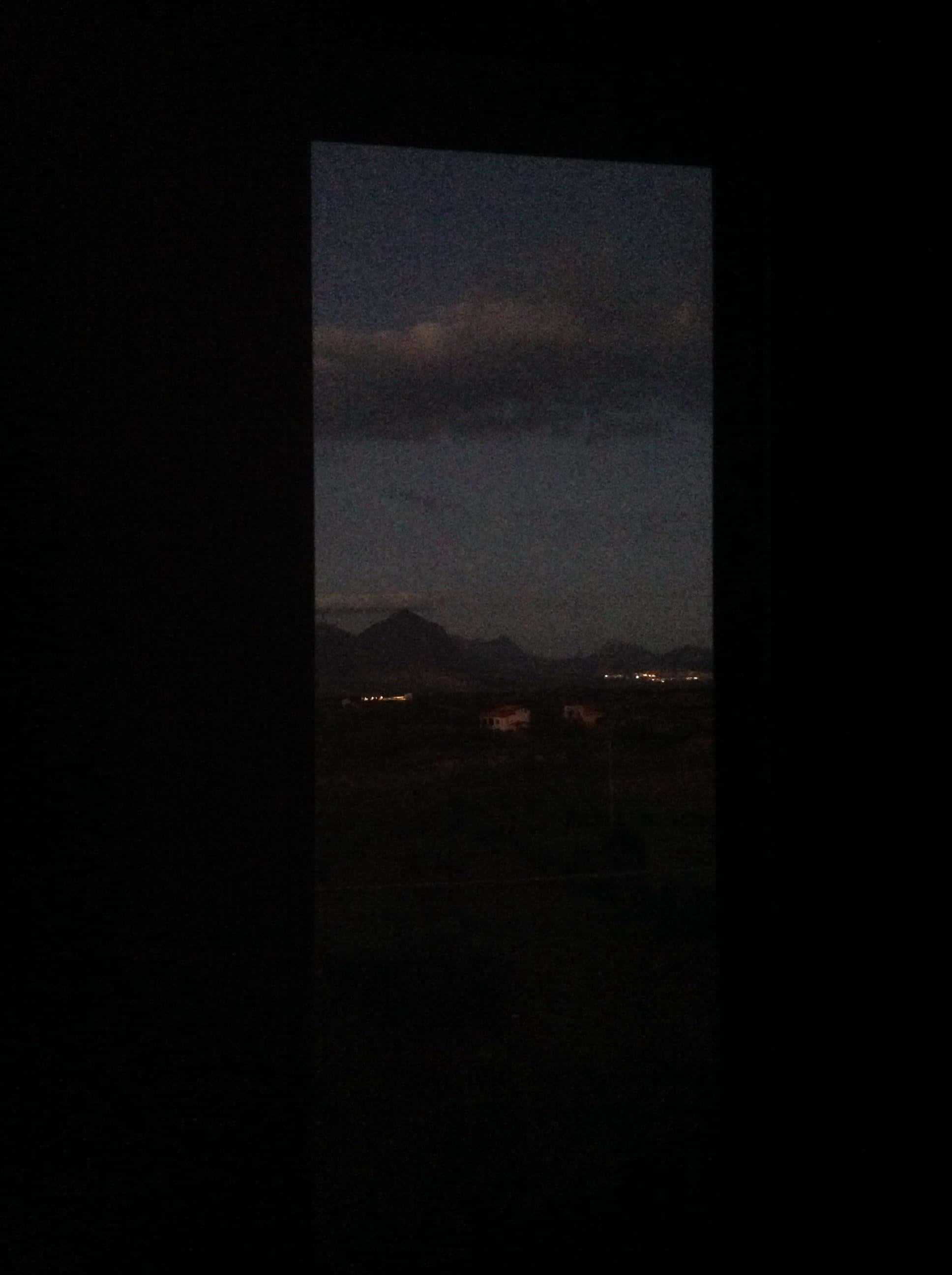 Aesthetic Black Grunge 桌布 壁紙 — 一扇昏暗的窗戶框住了多雲天空下遠山和散落村莊燈光的暮色景色。