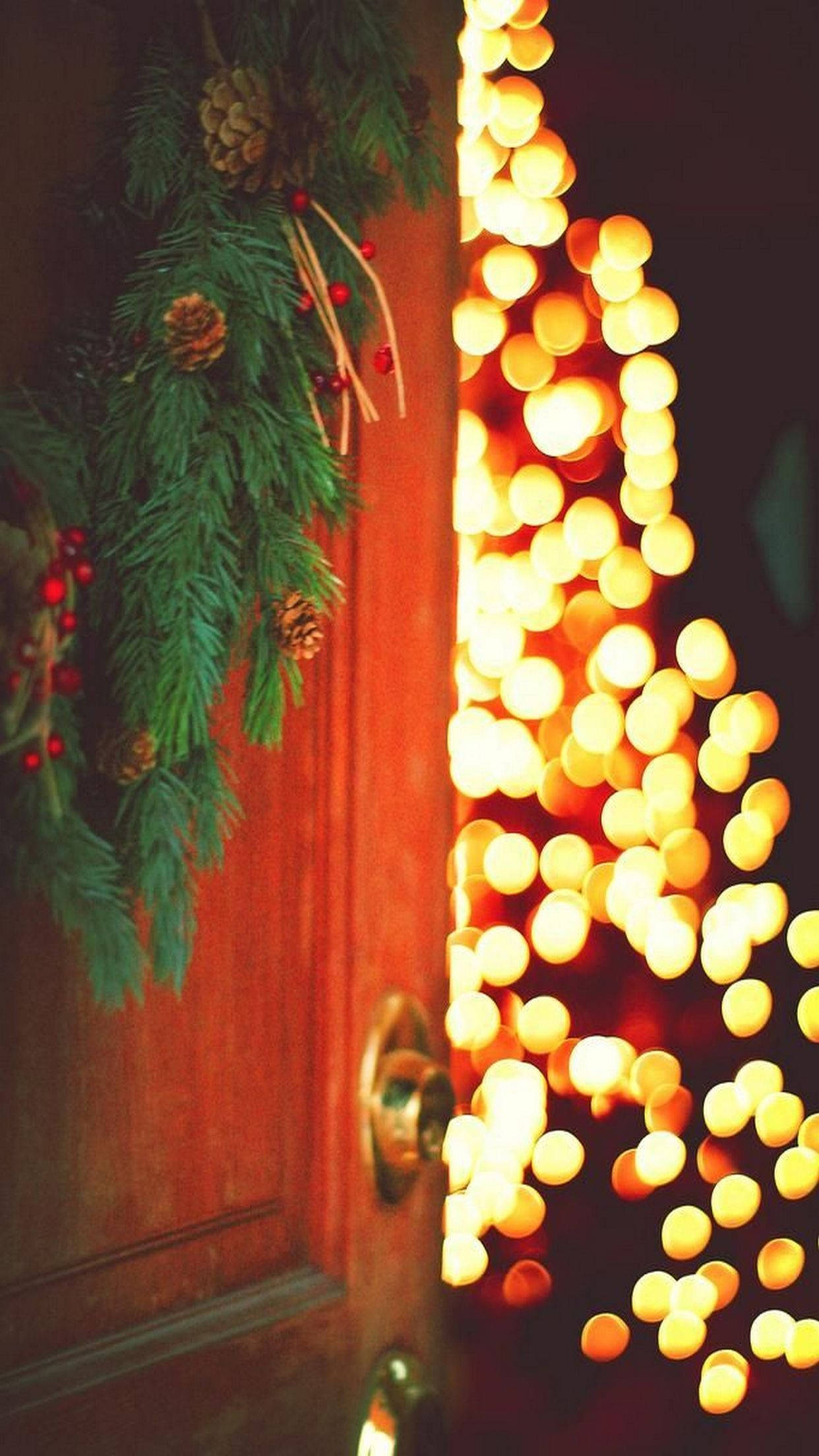 Christmas Lights iPhone 桌布 壁紙 — 一扇木門裝飾著松樹花環和松果，在模糊的金色仙女燈串旁溫暖地發光。
