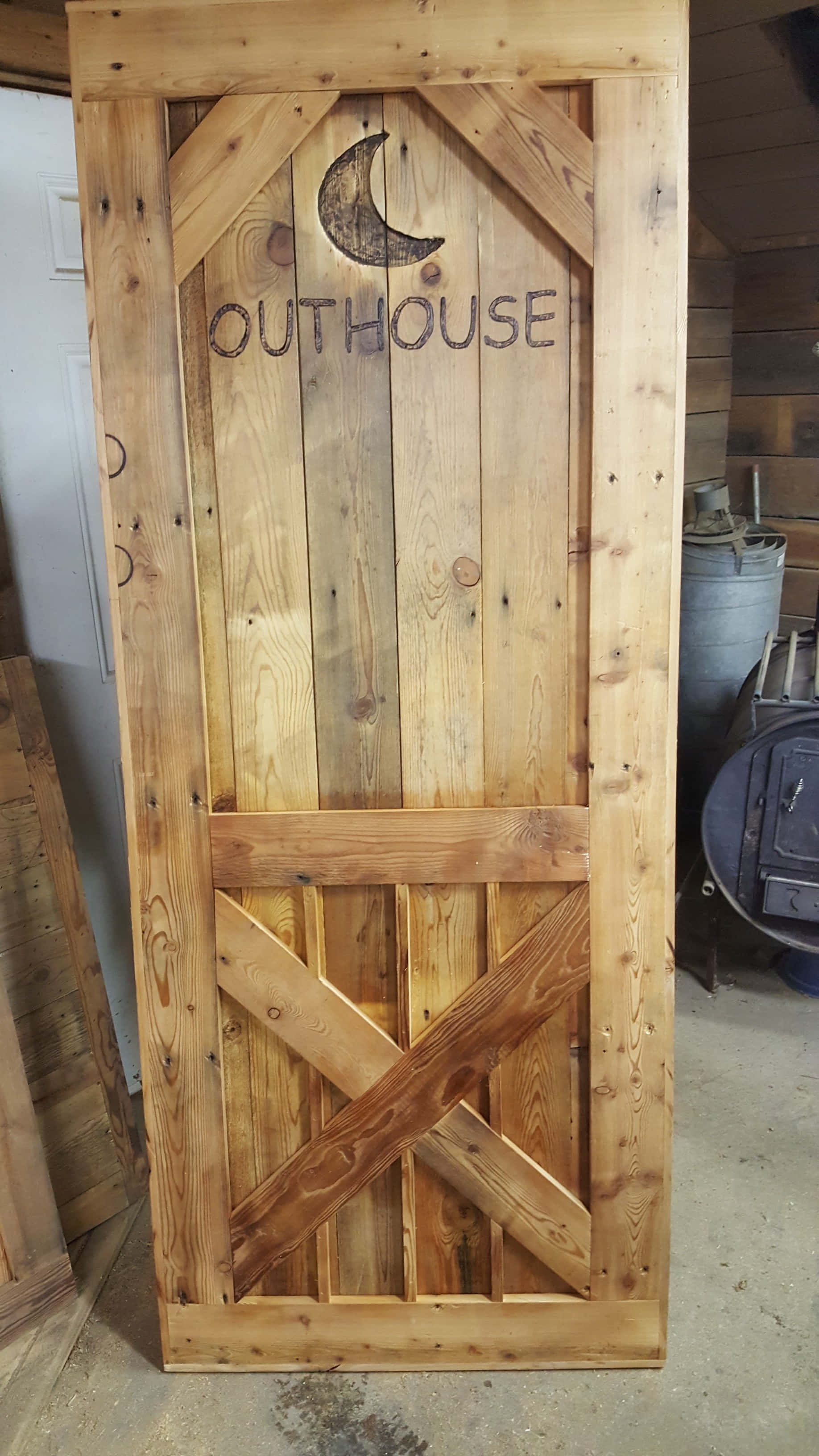 Outhouse 桌布 壁紙 — 一扇質樸的戶外廁所木門，上面刻有新月圖案和「OUTHOUSE」字樣，豎立在雜亂的工作室中。