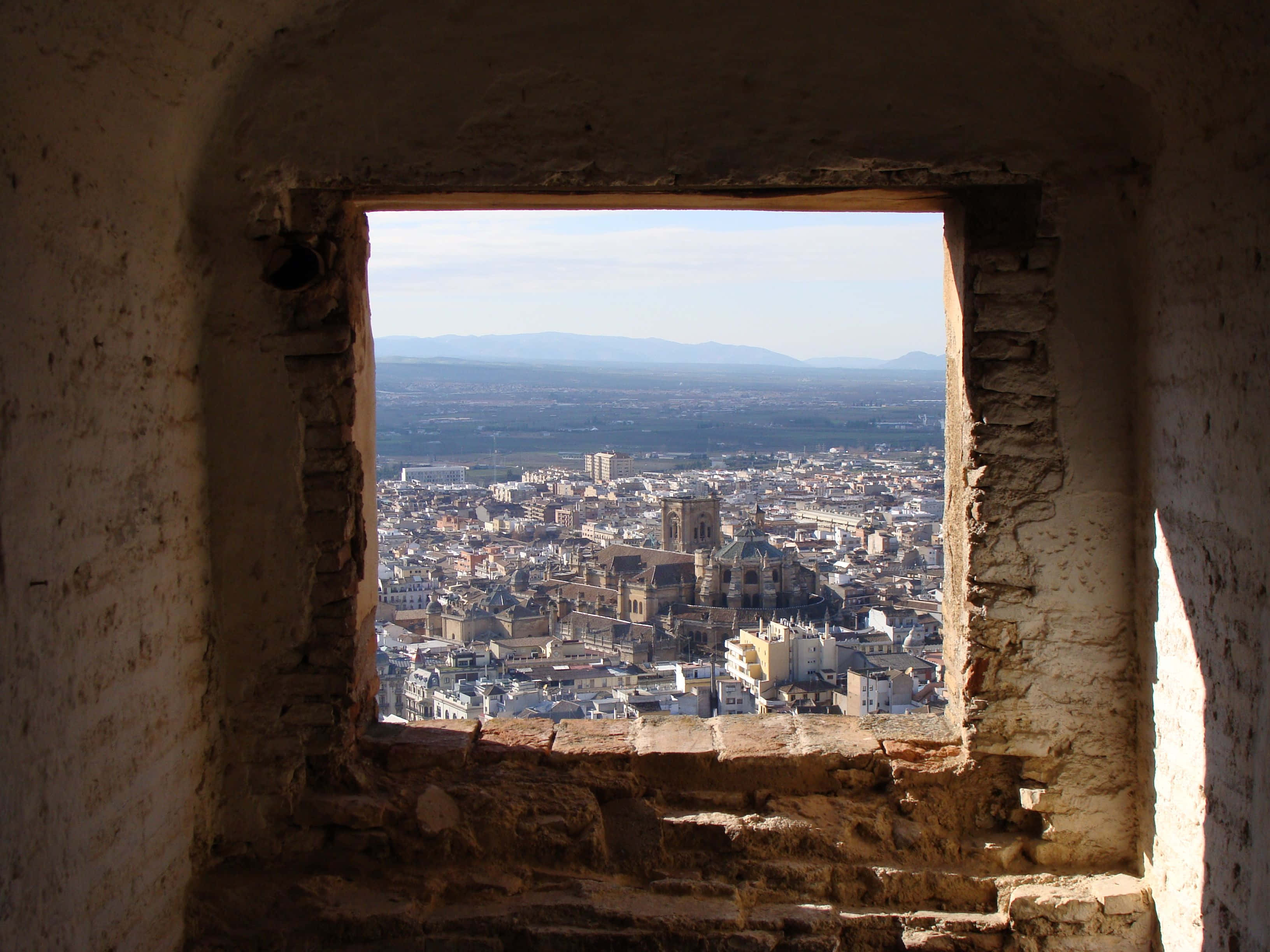 window 桌布 壁紙 — 一扇風化的石窗框住了陽光普照的城市景觀，歷史建築和遠處的山脈在晴朗的天空下延伸。