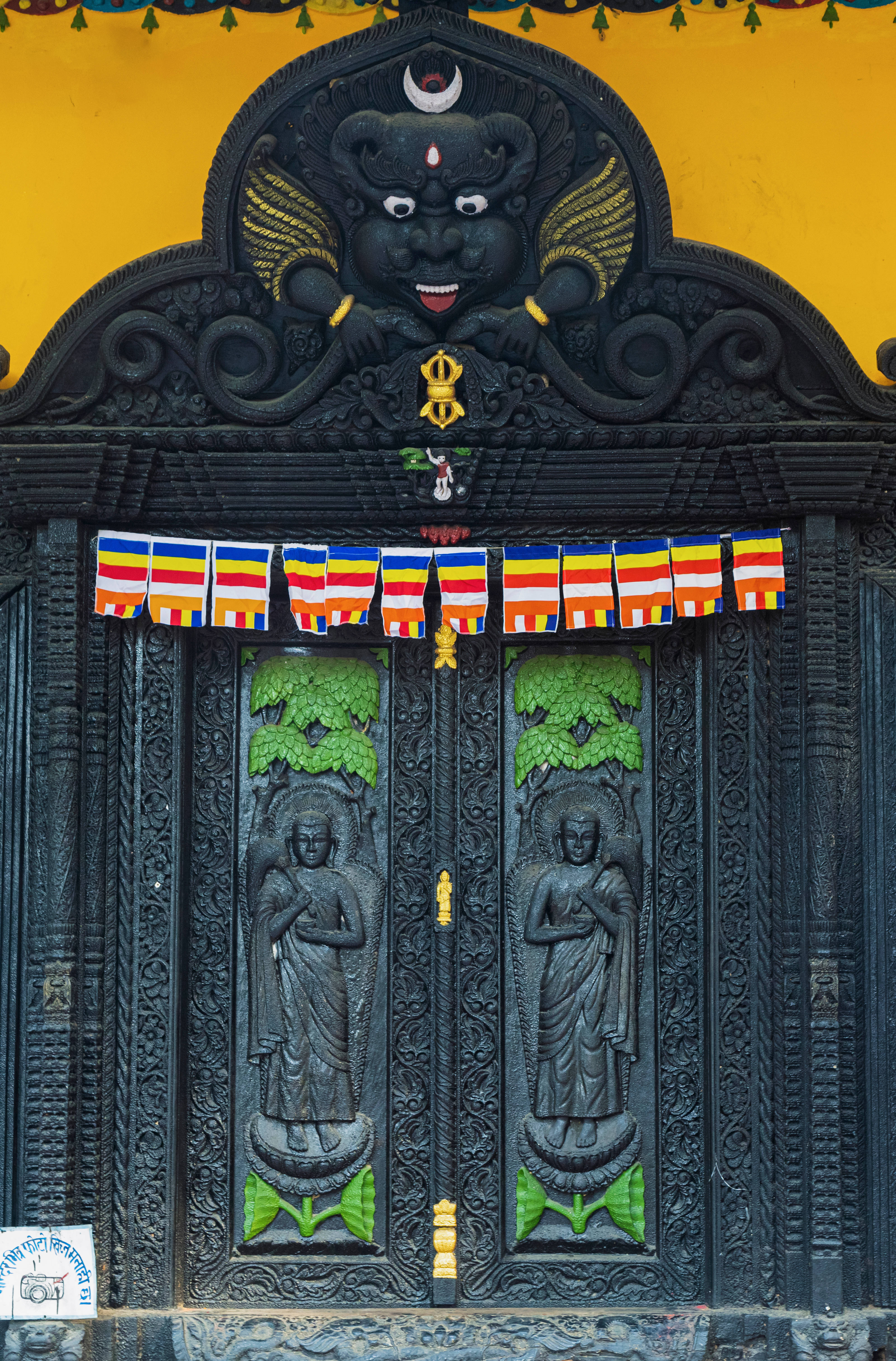 Nepal 桌布 壁紙 — 一扇黑暗華麗的寺廟門上雕刻著佛像，上方有一個兇猛的黑神，背景是鮮黃色的牆壁。