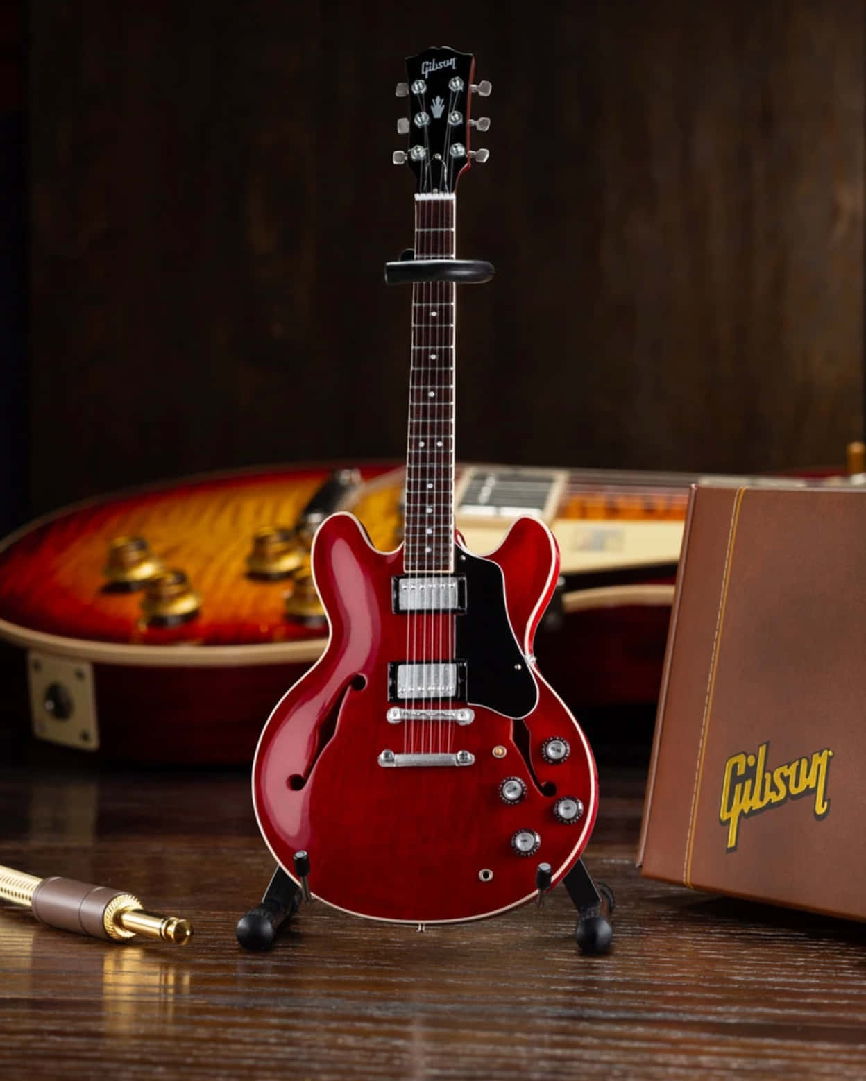 Guitar Aesthetic 桌布 壁紙 — 一把光澤的紅色Gibson吉他直立於木質表面上，旁邊有一個復古吉他盒和一條線纜。