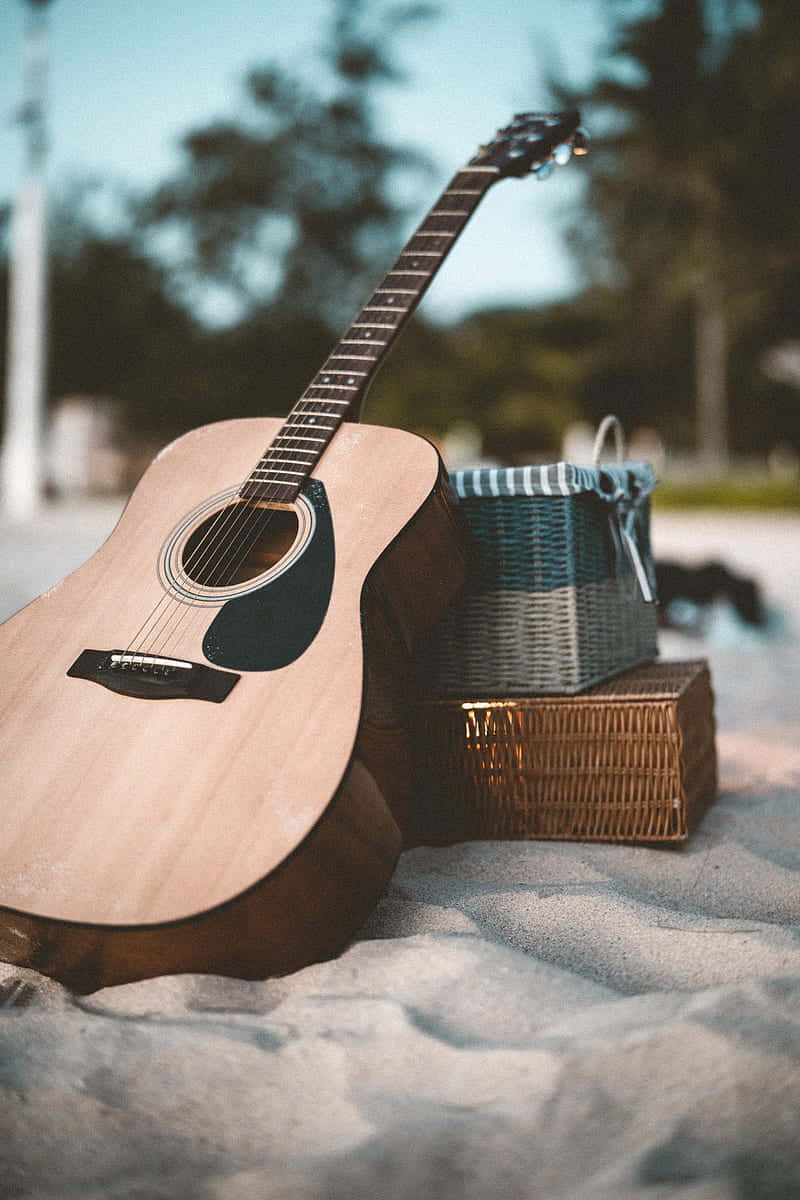 Acoustic Guitar 桌布 壁紙 — 一把原聲吉他靠在沙灘軟沙上的柳條野餐籃旁，天空晴朗。