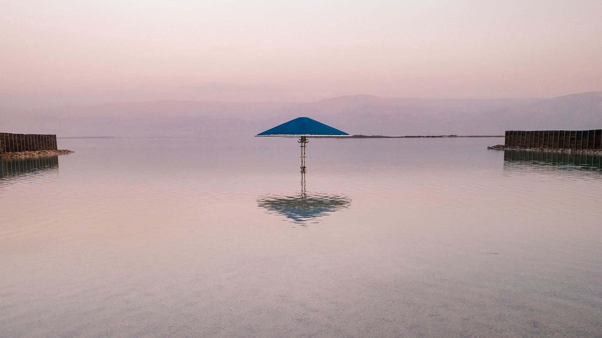 Beach Umbrella 桌布 壁紙 — 一把孤獨的藍色雨傘豎立在平靜的粉紅色調水中，遠山在柔和的暮色天空下。