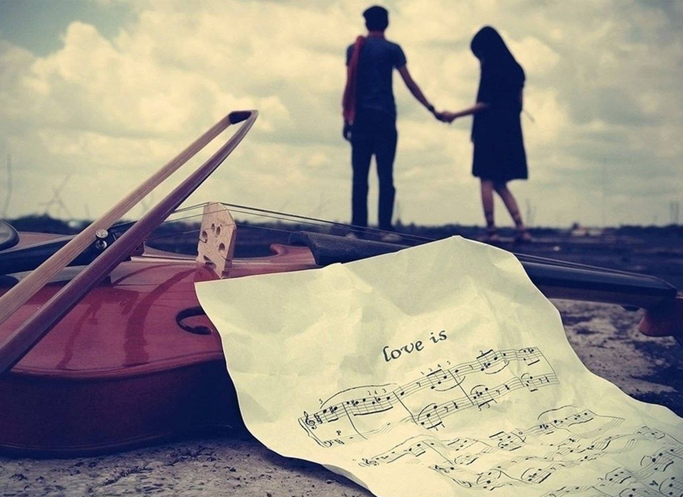 couple love 桌布 壁紙 — 一把小提琴和標題為「Love is」的樂譜放在前景，而一對情侶在遠處的山丘上手牽手，天空多雲。