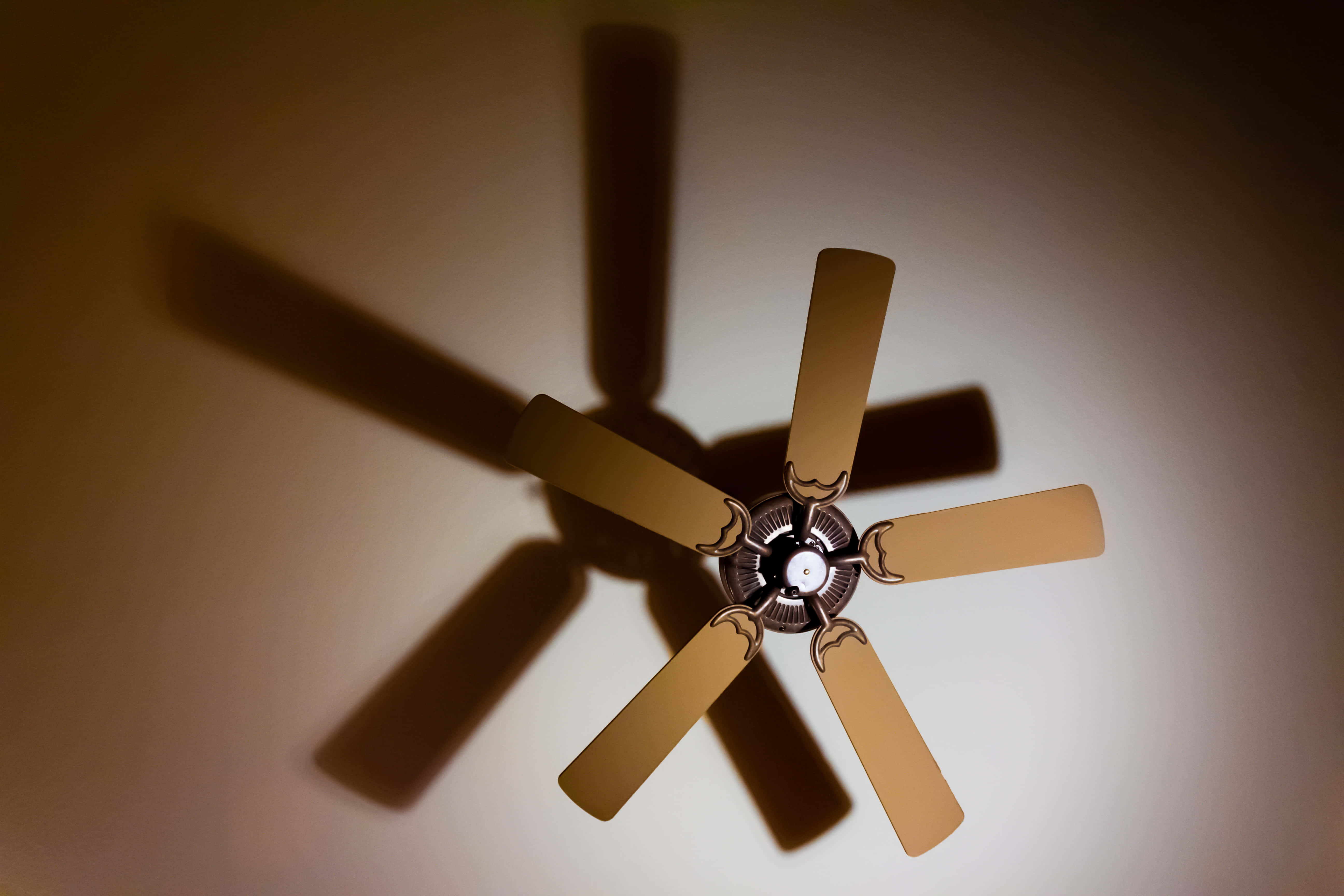 fan 桌布 壁紙 — 一把帶有木質葉片的吊扇在柔和的室內燈光下，在溫暖的米色牆壁上投下長長的陰影。