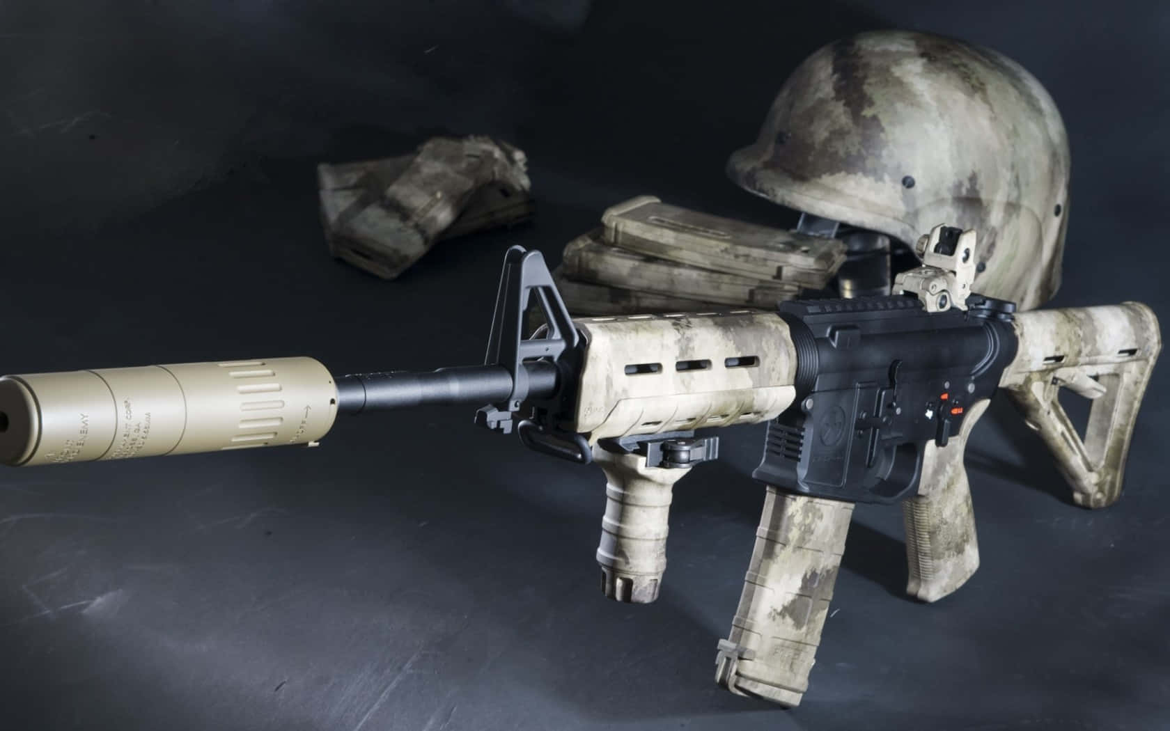 Rifle 桌布 壁紙 — 一把帶有消音器和頭盔的迷彩突擊步槍放在黑暗的表面上，附近有彈藥袋。