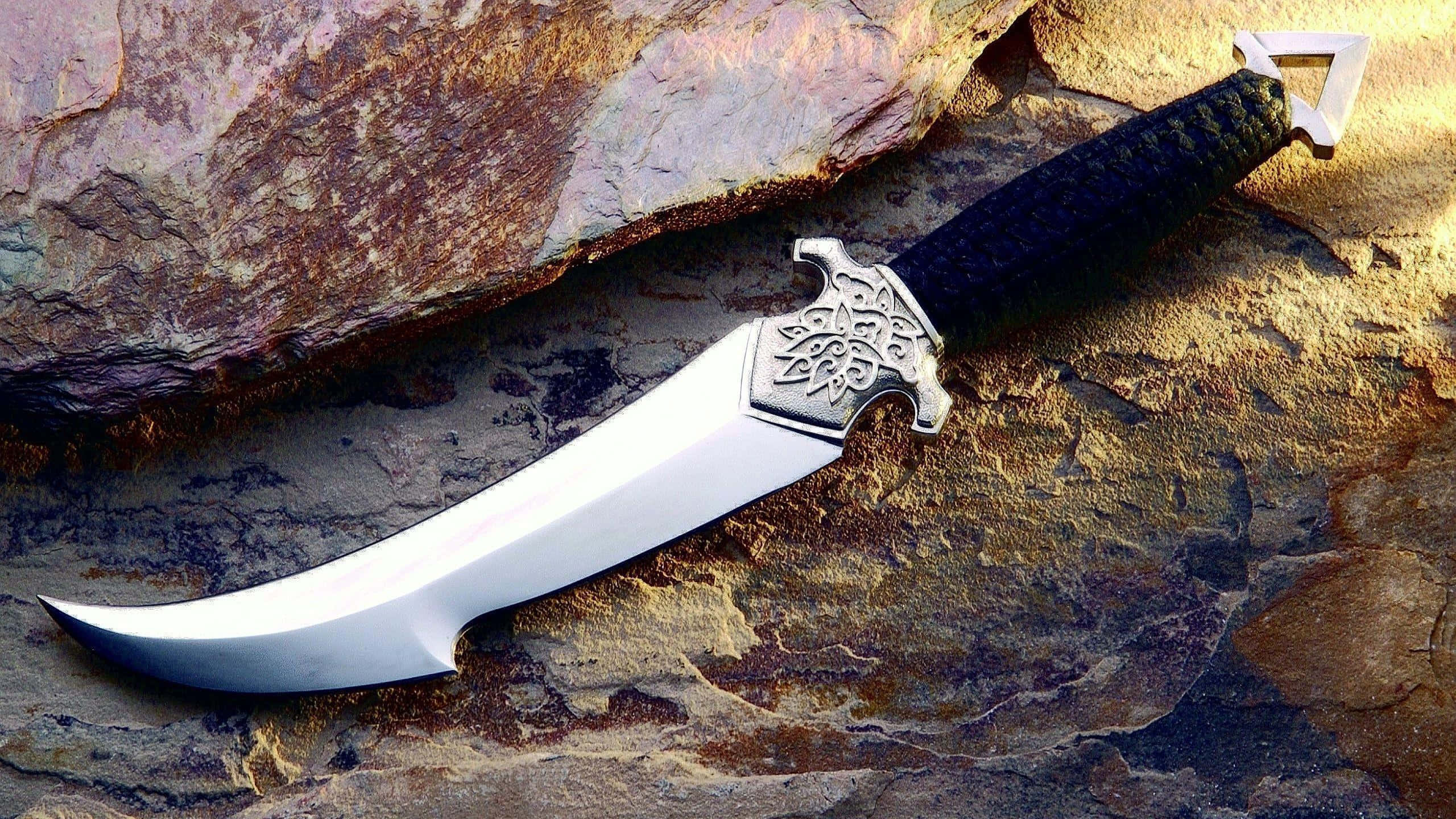 Knife 桌布 壁紙 — 一把彎曲的裝飾刀，帶有黑色紋理手柄，擱在堅固的陽光照射的岩石上。