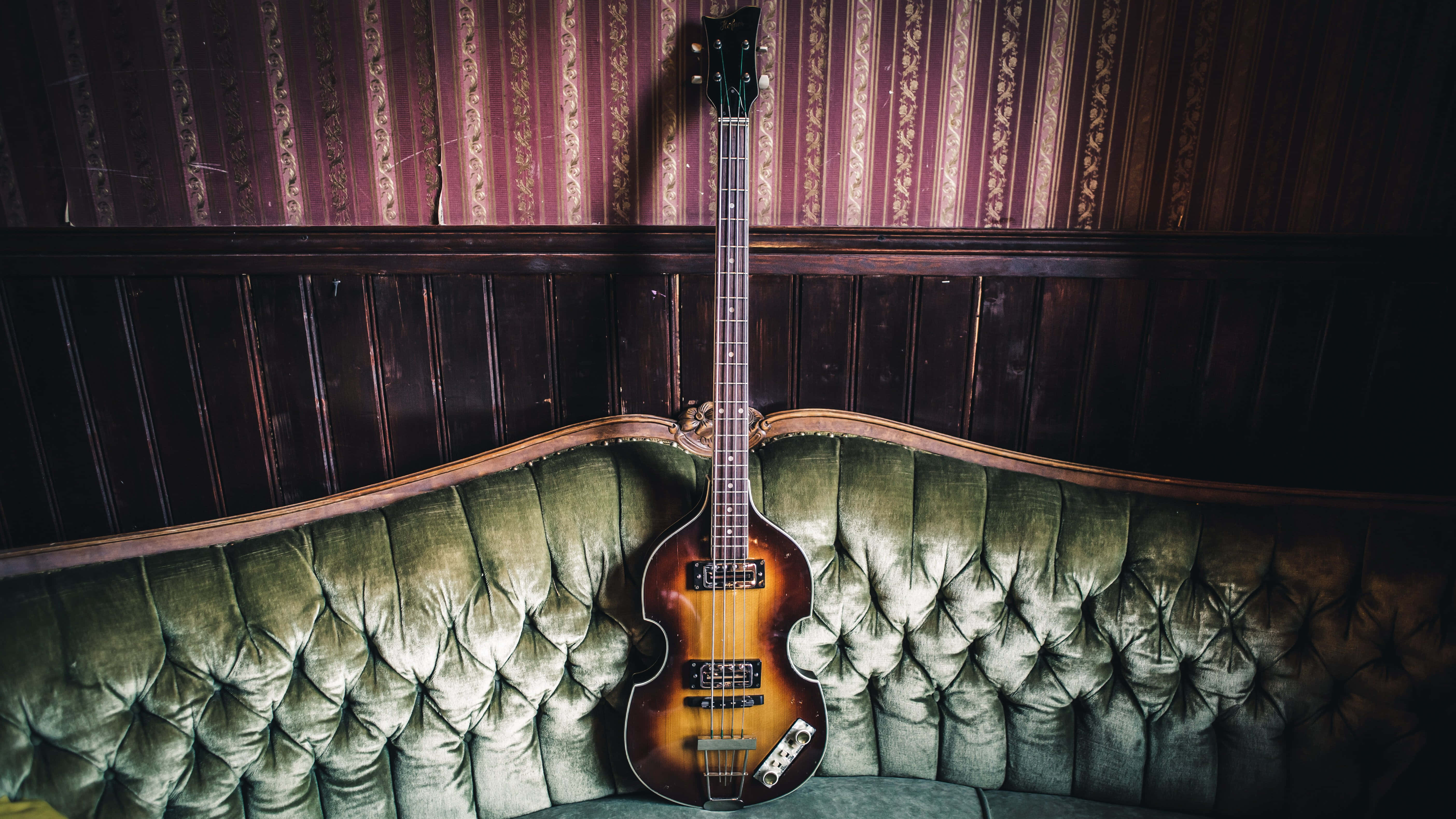 Bass Guitar 桌布 壁紙 — 一把復古旭日紋低音吉他優雅地放在一張簇絨綠色天鵝絨沙發上，背景是圖案豐富的勃艮第色壁紙。