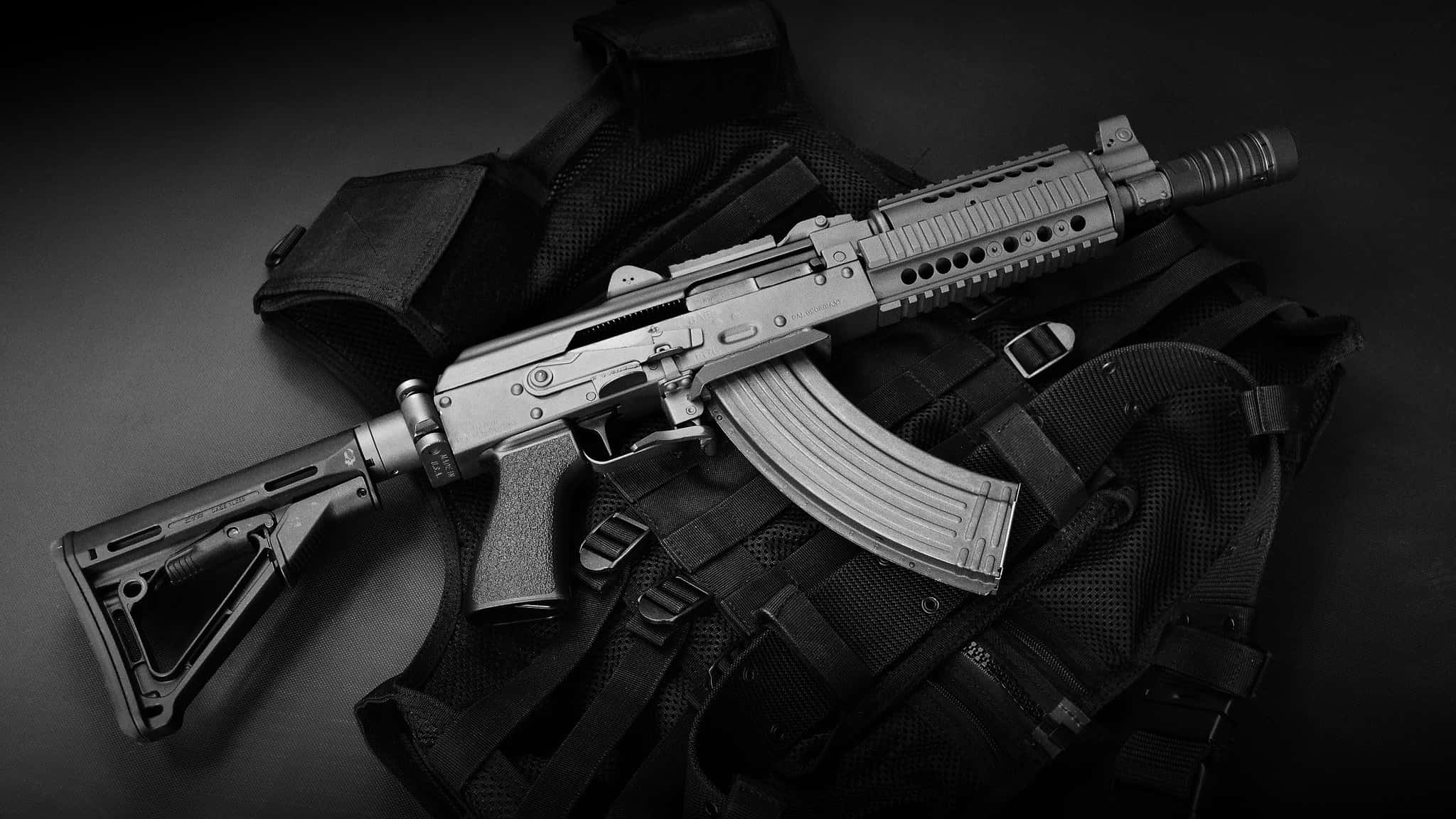 Rifle 桌布 壁紙 — 一把時尚的突擊步槍以對角線方式放在戰術背心上，背景為深色漸變，呈現高對比度的黑白效果。