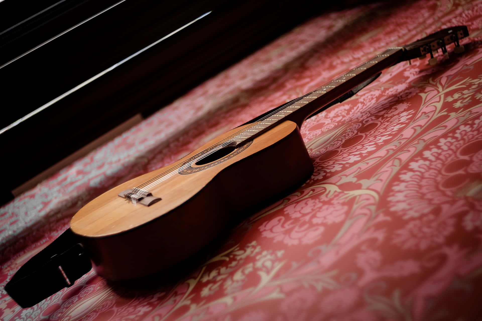 Guitar Aesthetic 桌布 壁紙 — 一把木製原聲吉他斜放在華麗的紅色錦緞圖案布料上，在溫暖的方向性燈光下。