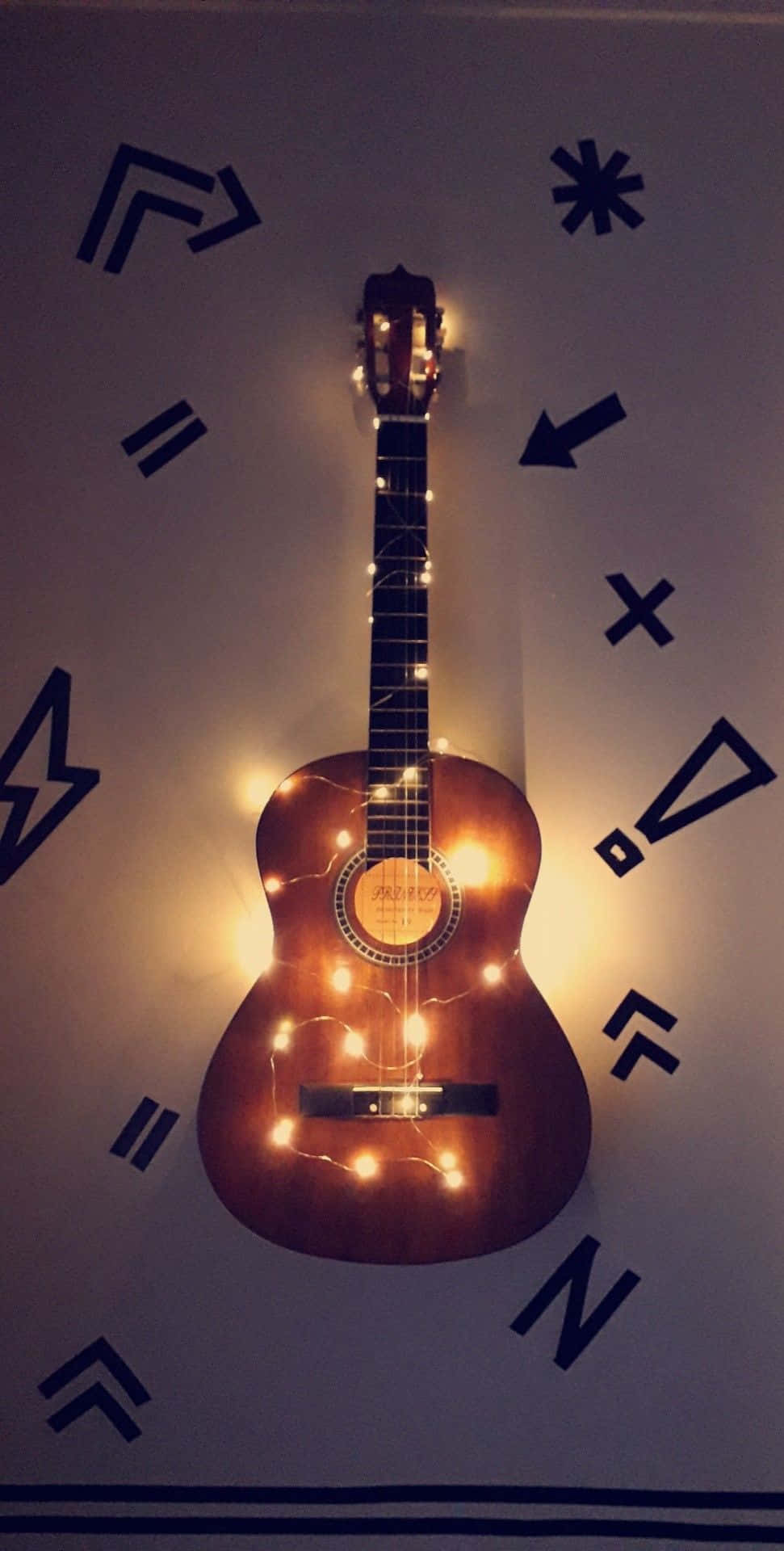 Guitar Aesthetic 桌布 壁紙 — 一把木製原聲吉他裝飾著溫暖的串燈，掛在飾有大膽黑色幾何符號的牆上。