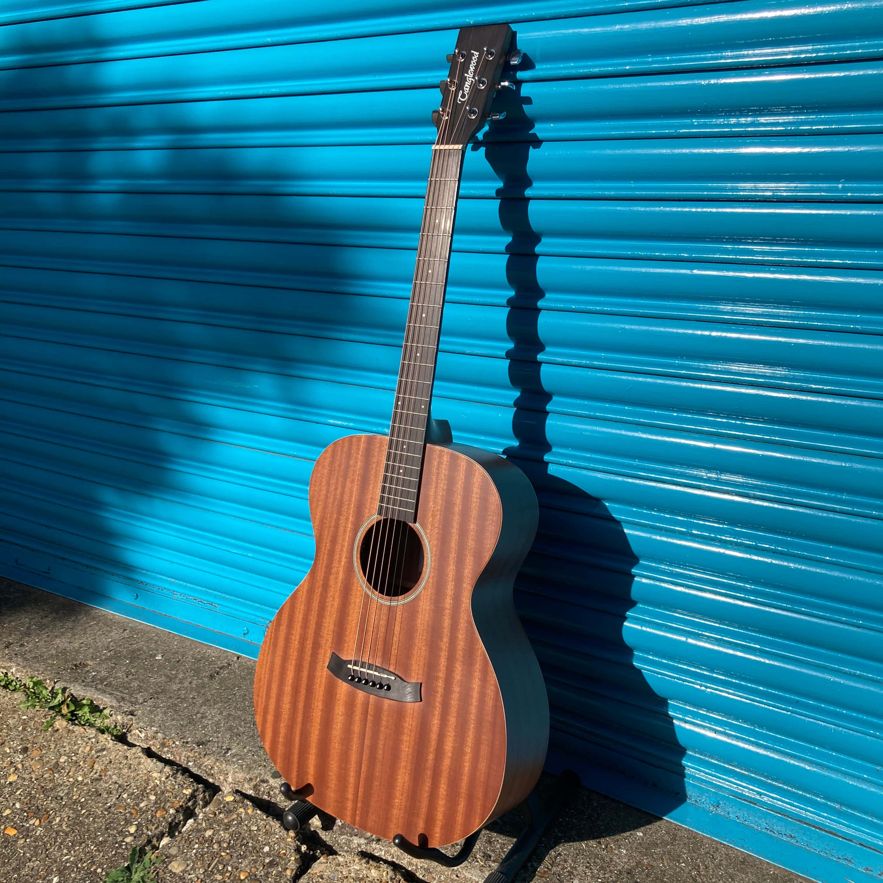Guitar Aesthetic 桌布 壁紙 — 一把木製原聲吉他靠在一扇亮藍色波紋金屬門上，在陽光下投下清晰的陰影。