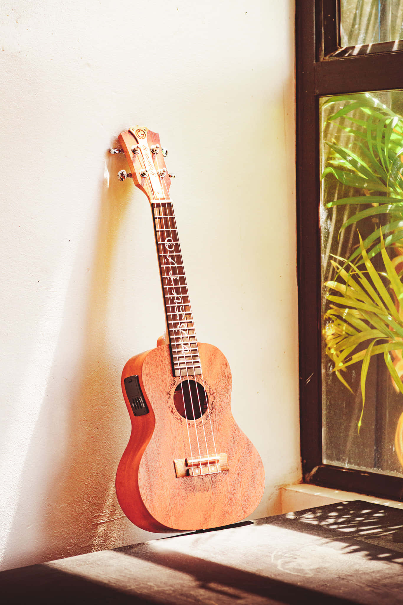 Guitar Aesthetic 桌布 壁紙 — 一把木製烏克麗麗靠在陽光照射的牆上，旁邊的窗戶有綠色樹葉在木質表面投下陰影。