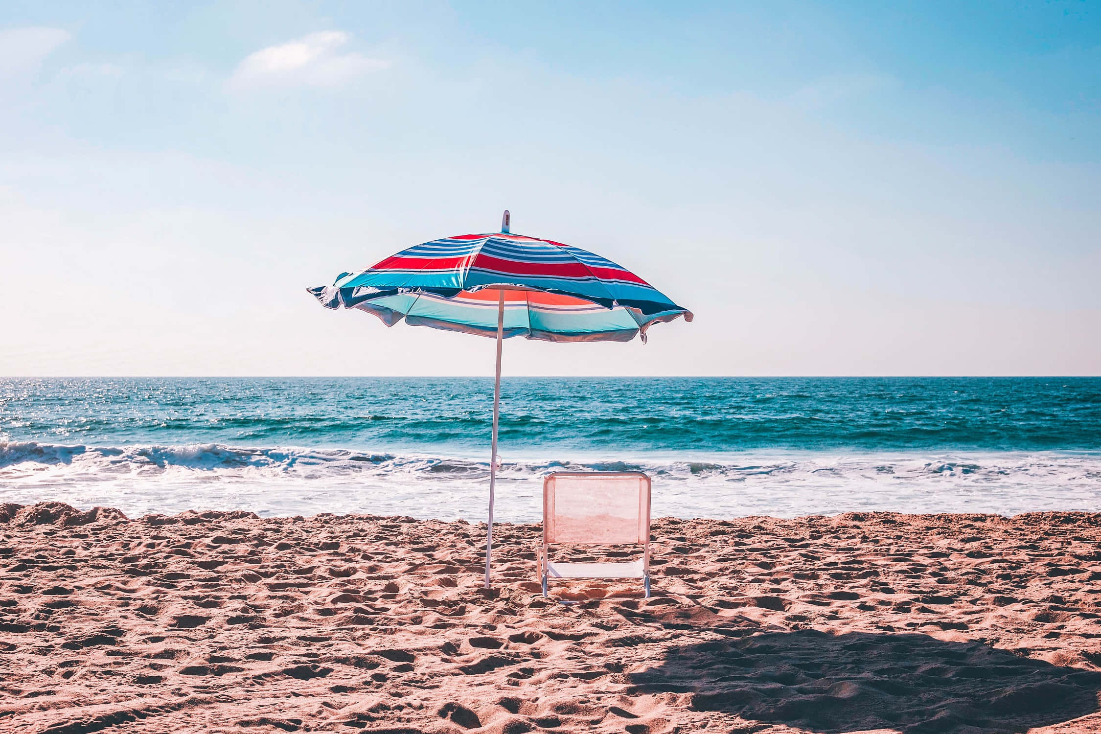 Beach Umbrella 桌布 壁紙 — 一把條紋沙灘傘和躺椅獨自放在陽光普照的沙灘上，面向湛藍的海洋，天空清澈蔚藍。