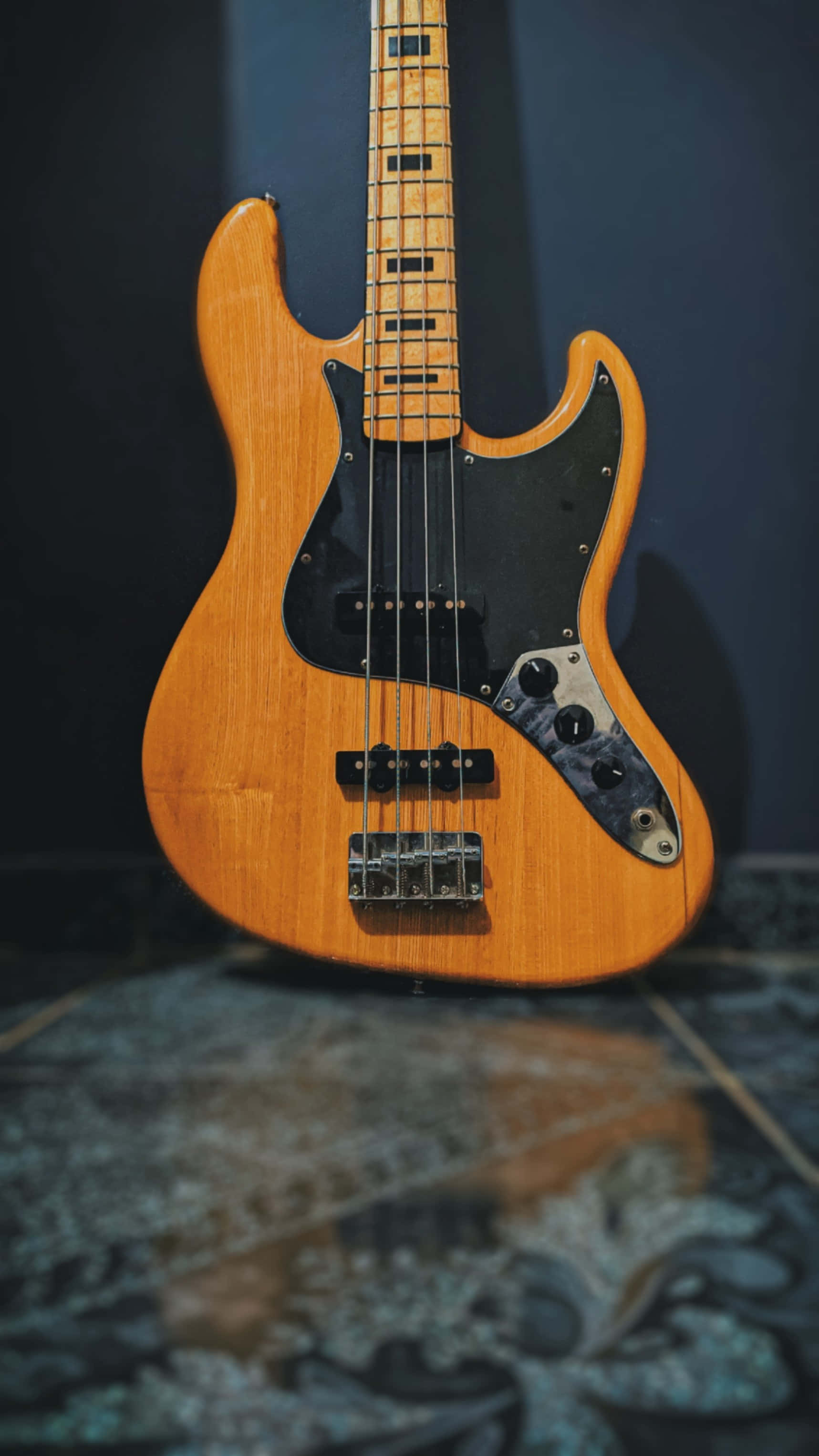 Bass Guitar 桌布 壁紙 — 一把漸層色飾面低音吉他直立靠著深色牆壁，放在圖案地毯上。